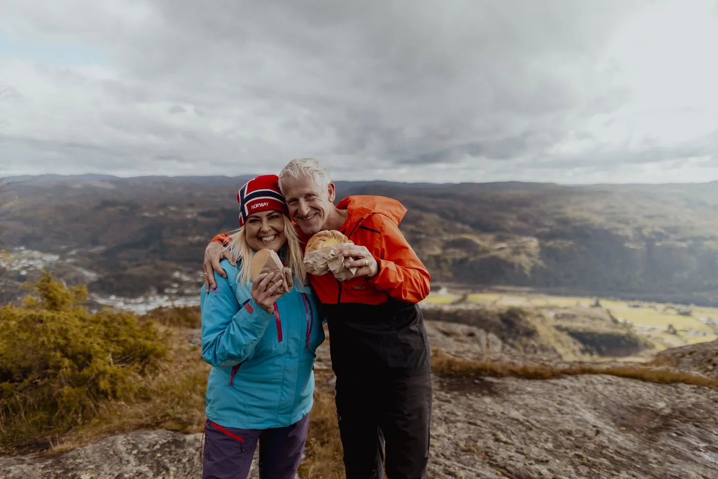 A couple in hiking clothes laughing at the camera on a rocky outcrop overlooking Kvinesdal in autumn colours
