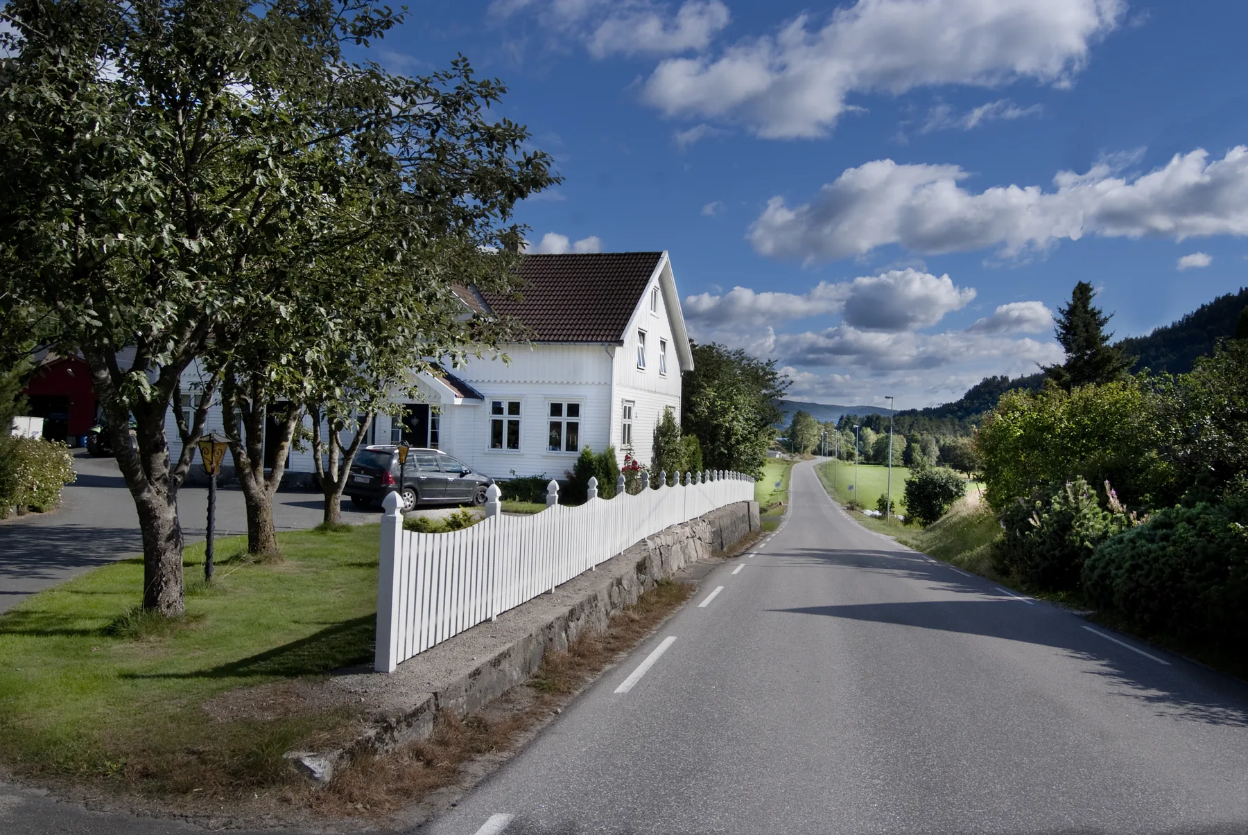 Hvitmalt trehus med stakittgjerde langs landeveien, og jorder videre nedover