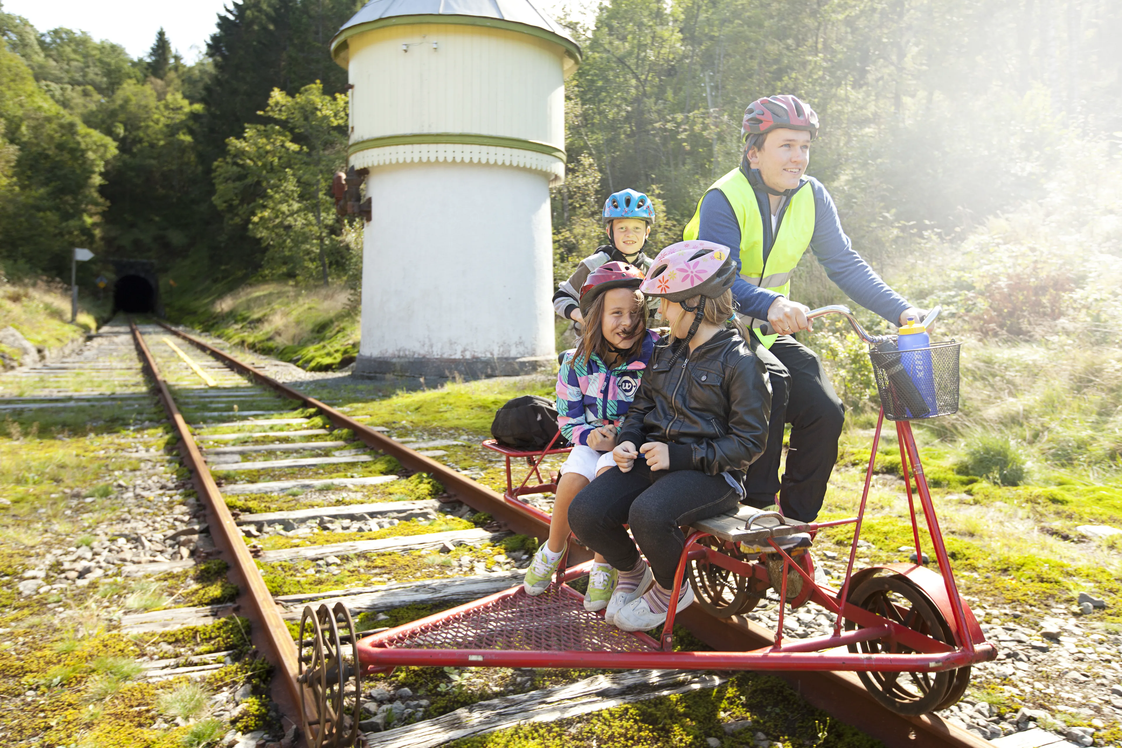 En voksen sykler en rød dresin med tre barn i hjelm om bord. Bak dem står et hvitt vanntårn med grønne lister og en tunnelmunning i skogen.