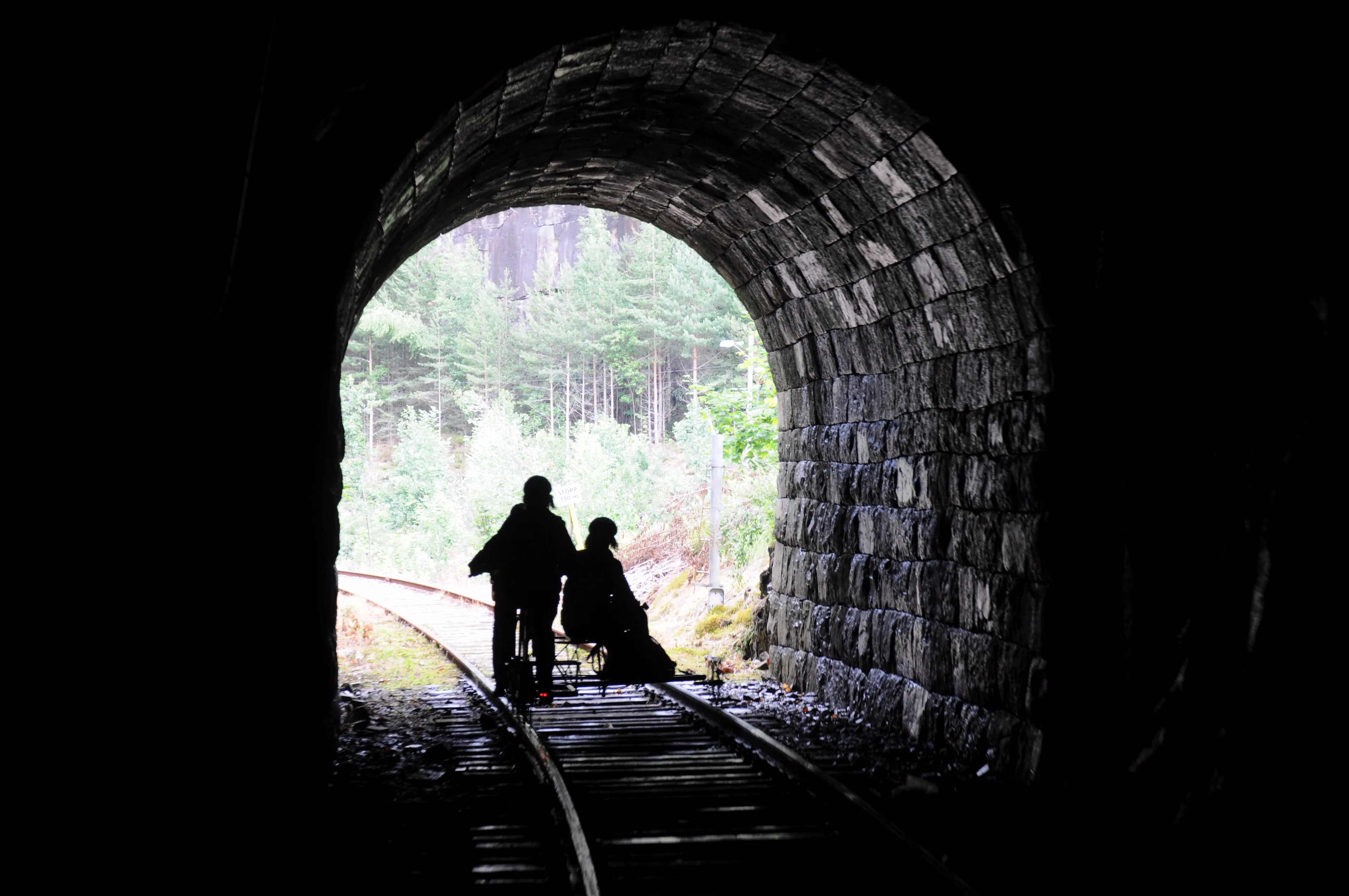 Inne i en tunnel med murt steinhvelv. To personer på dresin står i silhuett mot åpningen, der grønn skog lyser i dagslys.