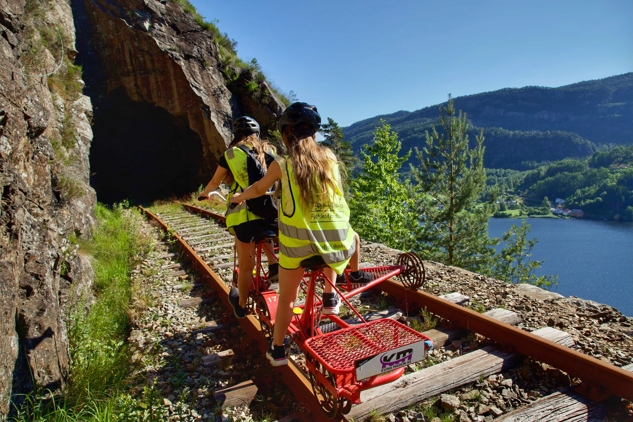 To jenter i refleksvest sykler en rød dresin mot en tunnelmunning i fjellet. Til høyre ligger fjorden og bygda i sol.