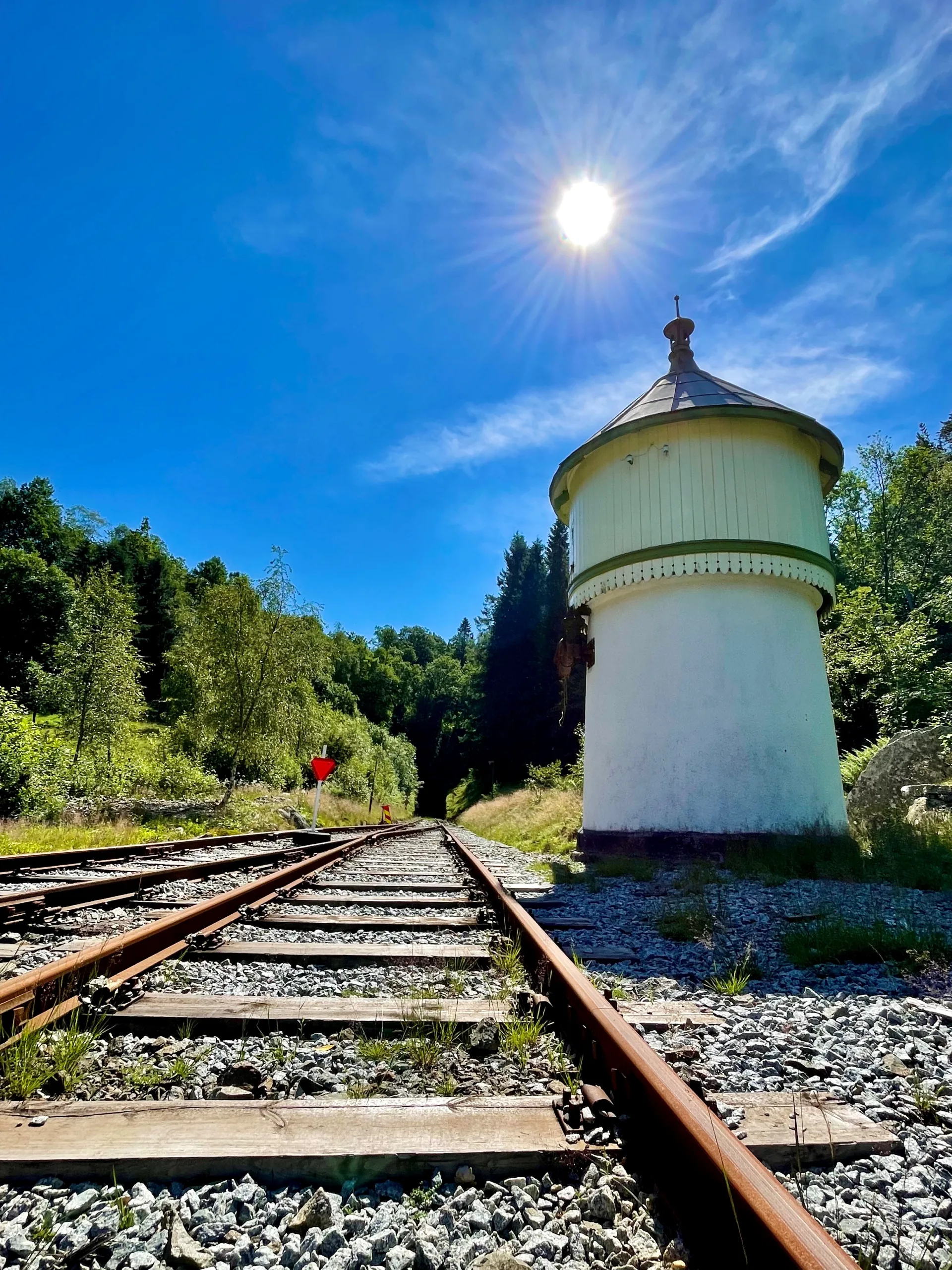 Et hvitt vanntårn med spisst tak ved siden av rustne skinner, fotografert nedenfra mot blå himmel og sterk sol.
