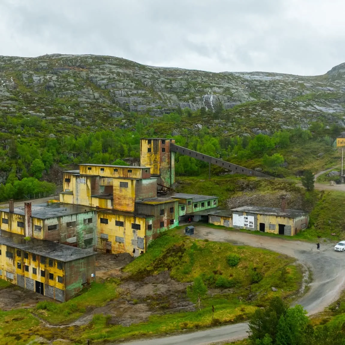 The mine works at Knaben seen from the air: yellow and green concrete buildings with peeling paint, a tall tower and a sloping conveyor up towards the mountainside. A gravel road curves past, and grey rocky peaks rise behind.