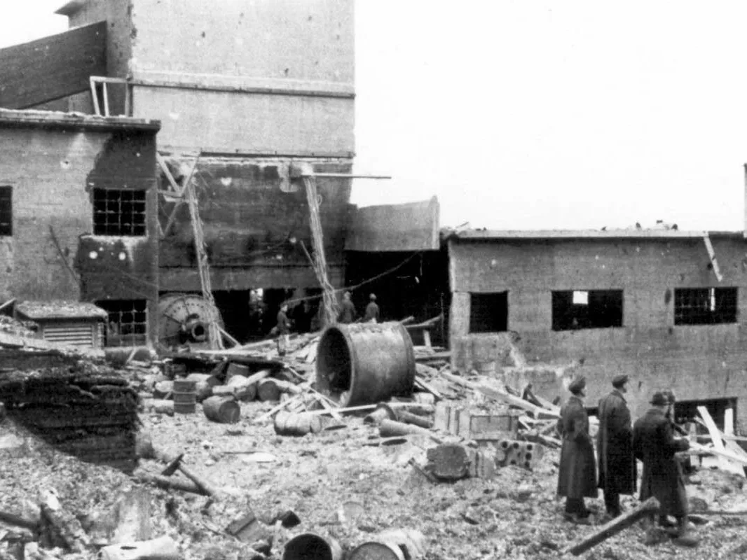 Black-and-white photograph: damaged concrete buildings with broken windows and a collapsed roof. The ground is strewn with barrels, beams and rubble. Three men in long coats stand looking on.