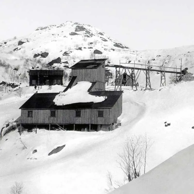Black-and-white photograph: wooden mine buildings in deep snow below a mountain peak. A conveyor on wooden trestles runs from the buildings up the mountainside.