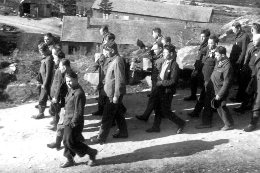 Black-and-white photograph: a large group of men in work clothes walking close together along a gravel road. Behind them are low wooden houses and a shed.