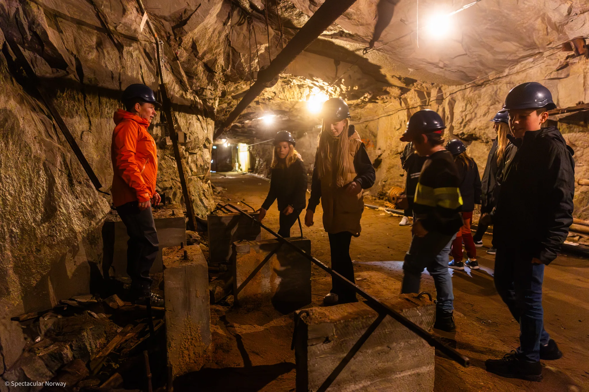 Inside the old mine: a guide in an orange jacket shows a group of children in helmets around a mine tunnel with rough rock walls, concrete foundations and lamps in the ceiling.
