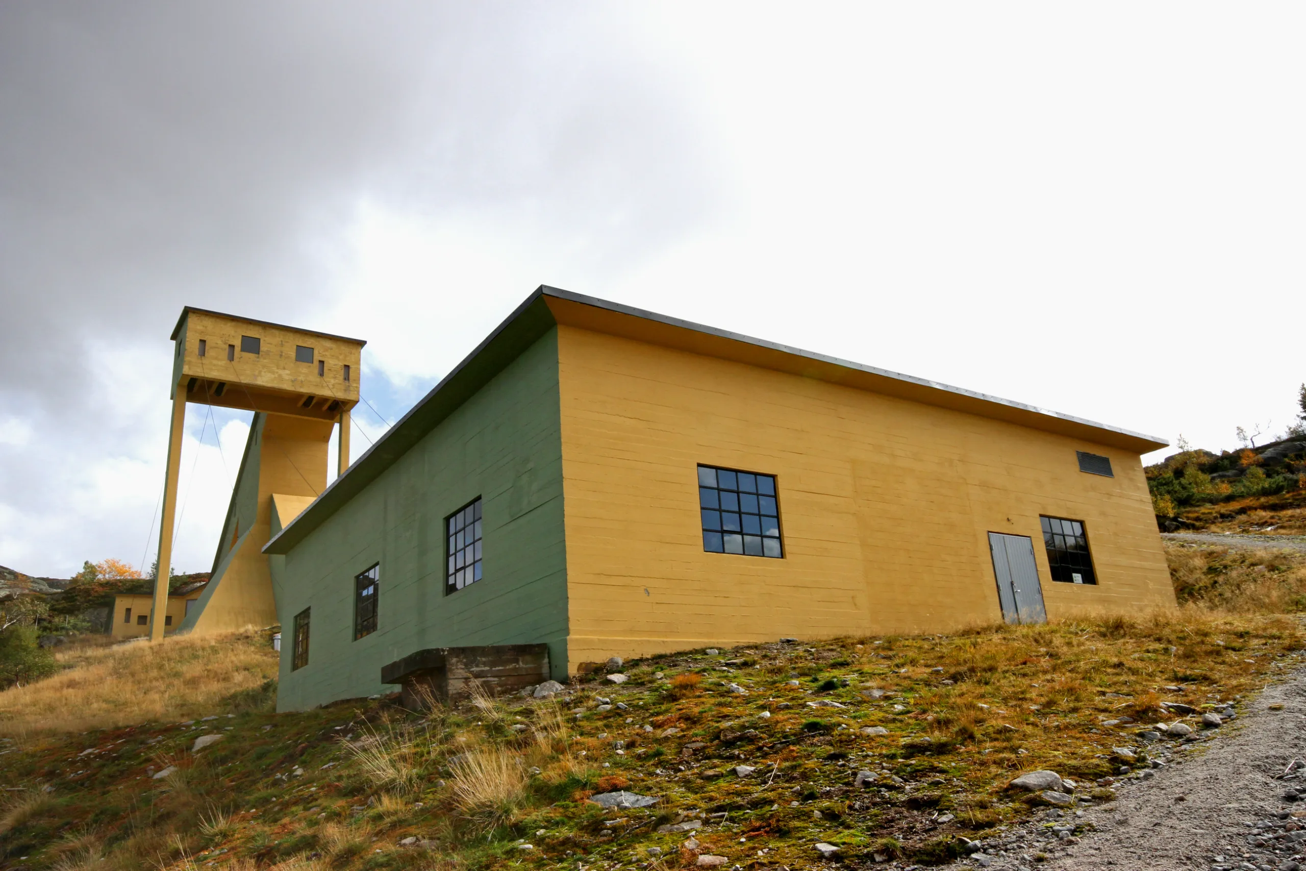 A long, low concrete building painted ochre and green, photographed from below on a heather-covered slope. Behind it a tall yellow tower rises against a grey sky.
