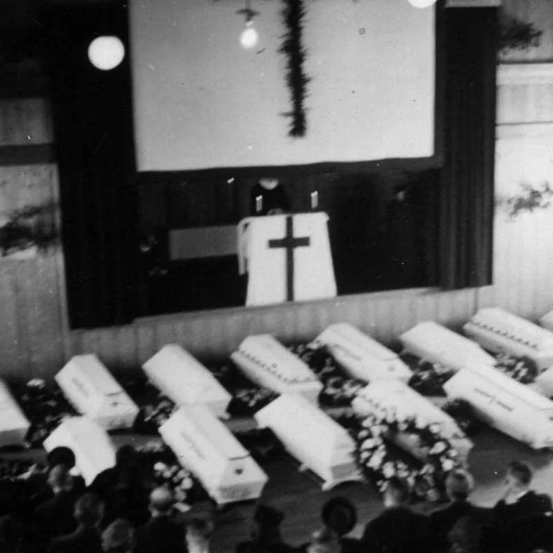 Black-and-white photograph: many white coffins with wreaths stand in a row in front of a stage with an altar and a cross. People are seated in the foreground.