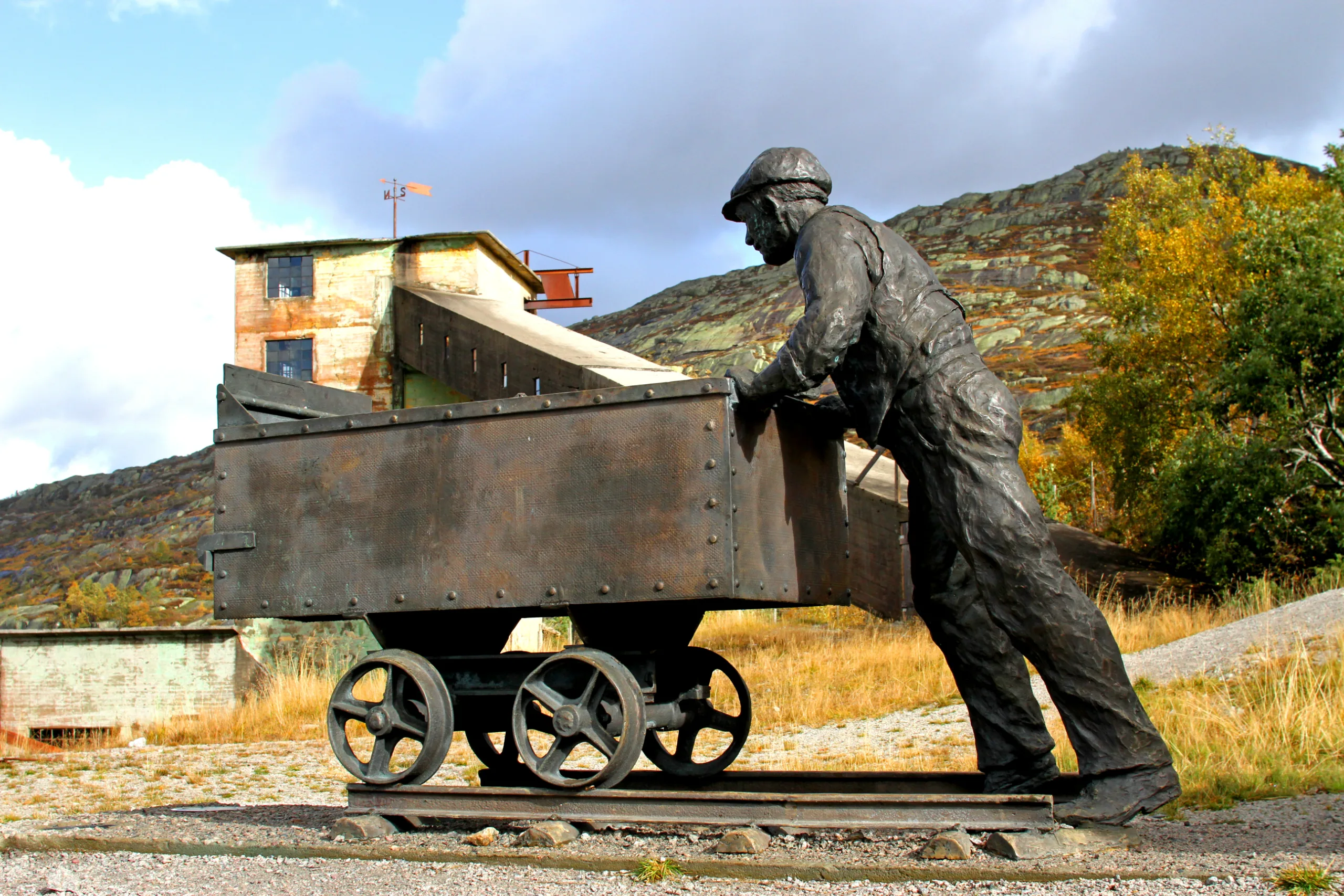 A bronze sculpture of a miner in a flat cap pushing a heavy ore cart on rails. Behind him stands an old mine tower with a weathervane, and a mountainside in autumn colours.