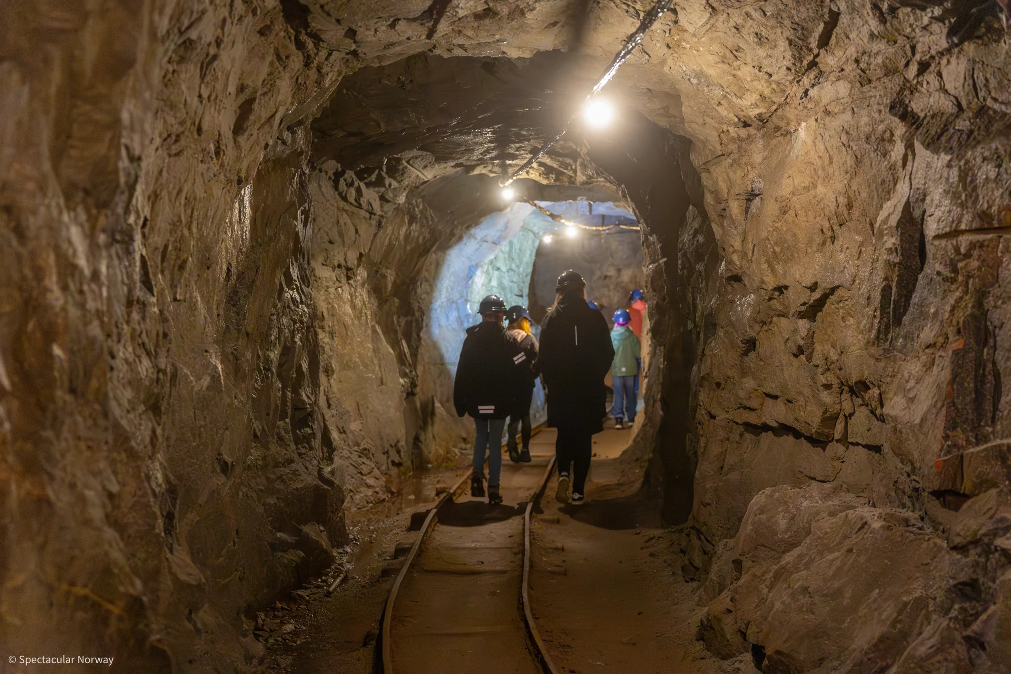 A group in helmets walking through a narrow, lit mine tunnel with rails along the floor.
