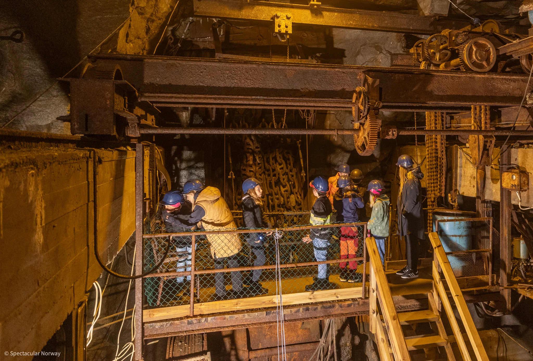 Children and adults in helmets stand on a railed platform among old machinery, cogwheels and beams deep inside the mine.
