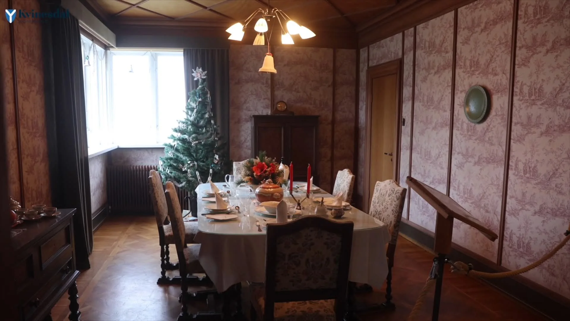 A dining room with a long laid table, high-backed chairs, a Christmas tree and a chandelier under floral wallpaper.