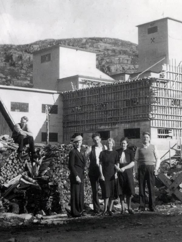 Black-and-white photograph: five young men and women in smart clothes stand in front of a large woodpile and mine buildings under construction. A sixth sits on top of the woodpile.