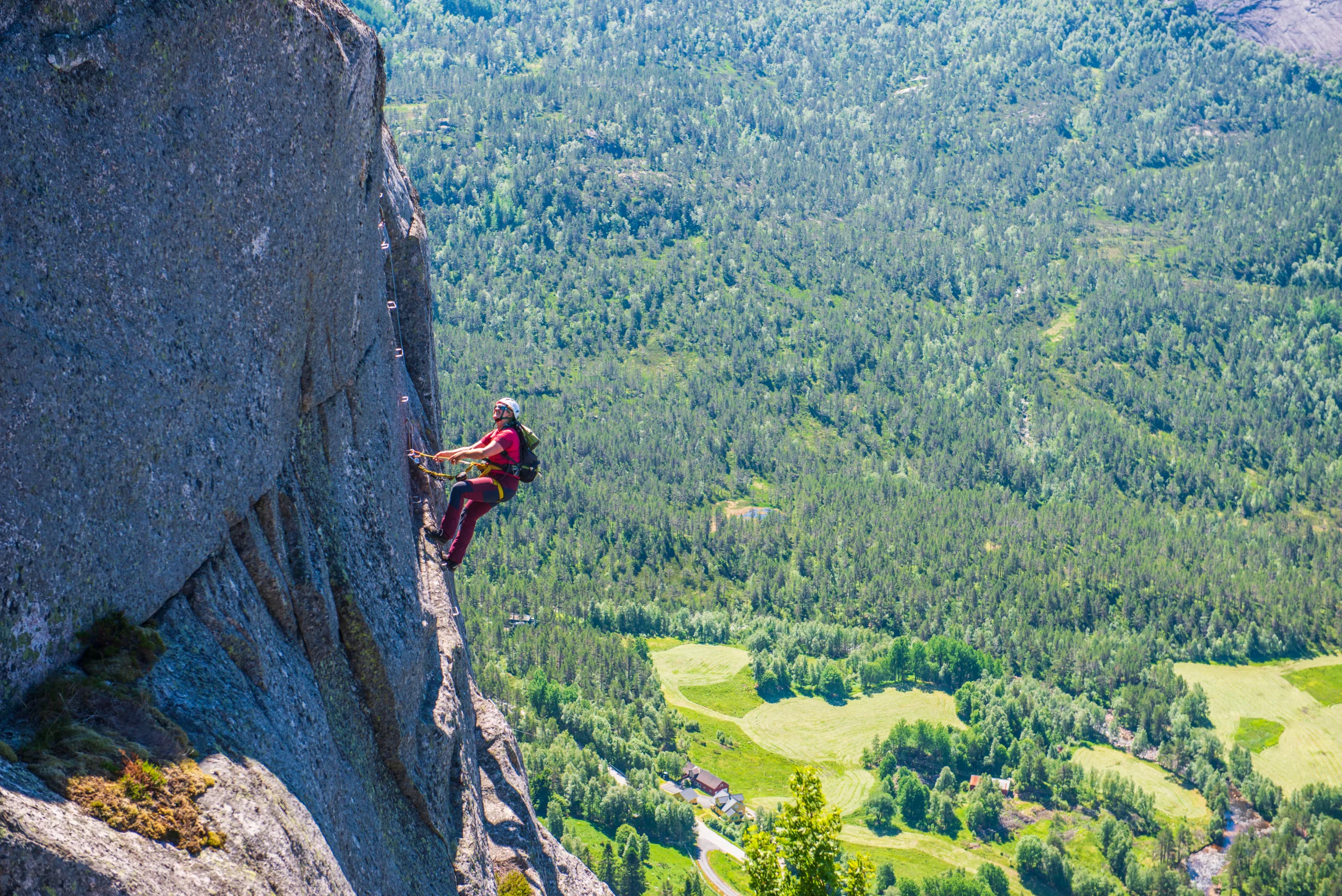A climber in a red T-shirt and helmet hangs secured to a cable on a vertical granite wall. Far below lies a wooded valley with fields and red farmhouses.