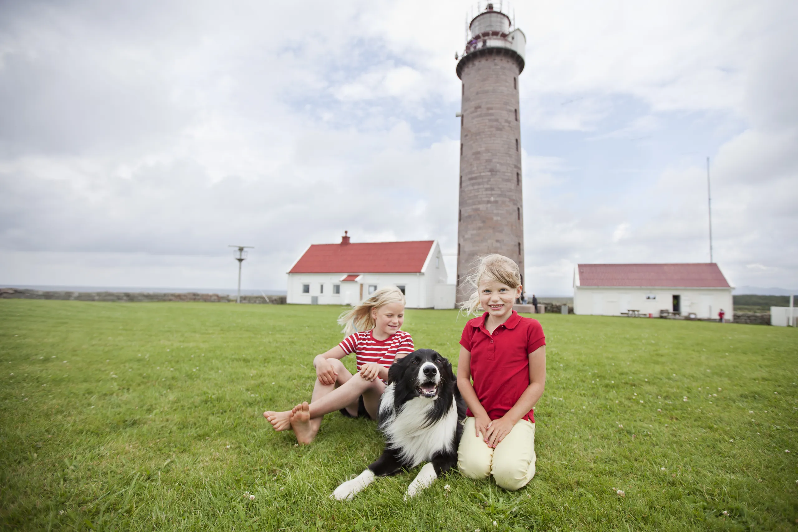 To jenter og en hund i gresset foran fyrtårnet og de hvite husene