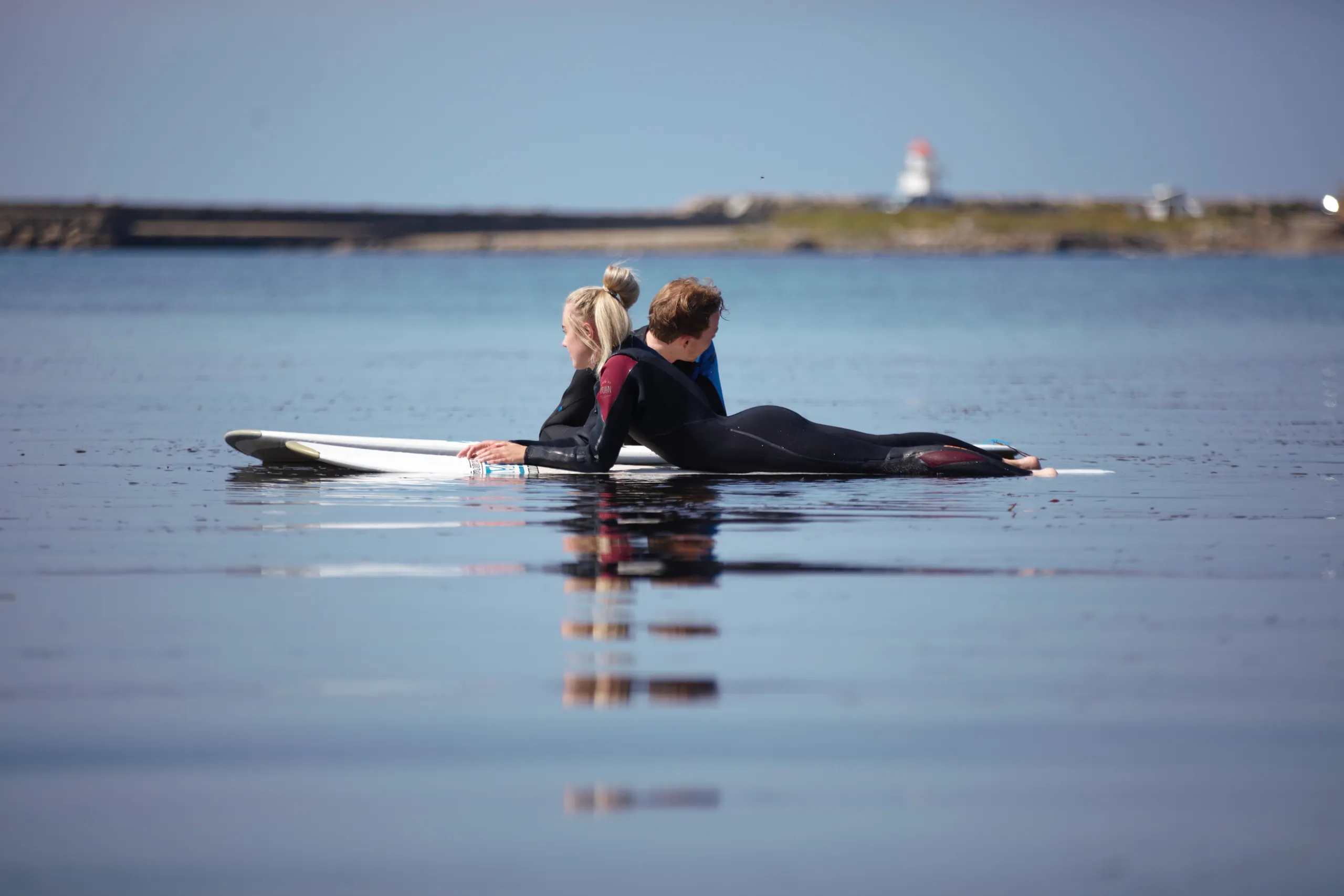 To unge surfere ligger på brettene i blikkstille vann, med et fyr i bakgrunnen