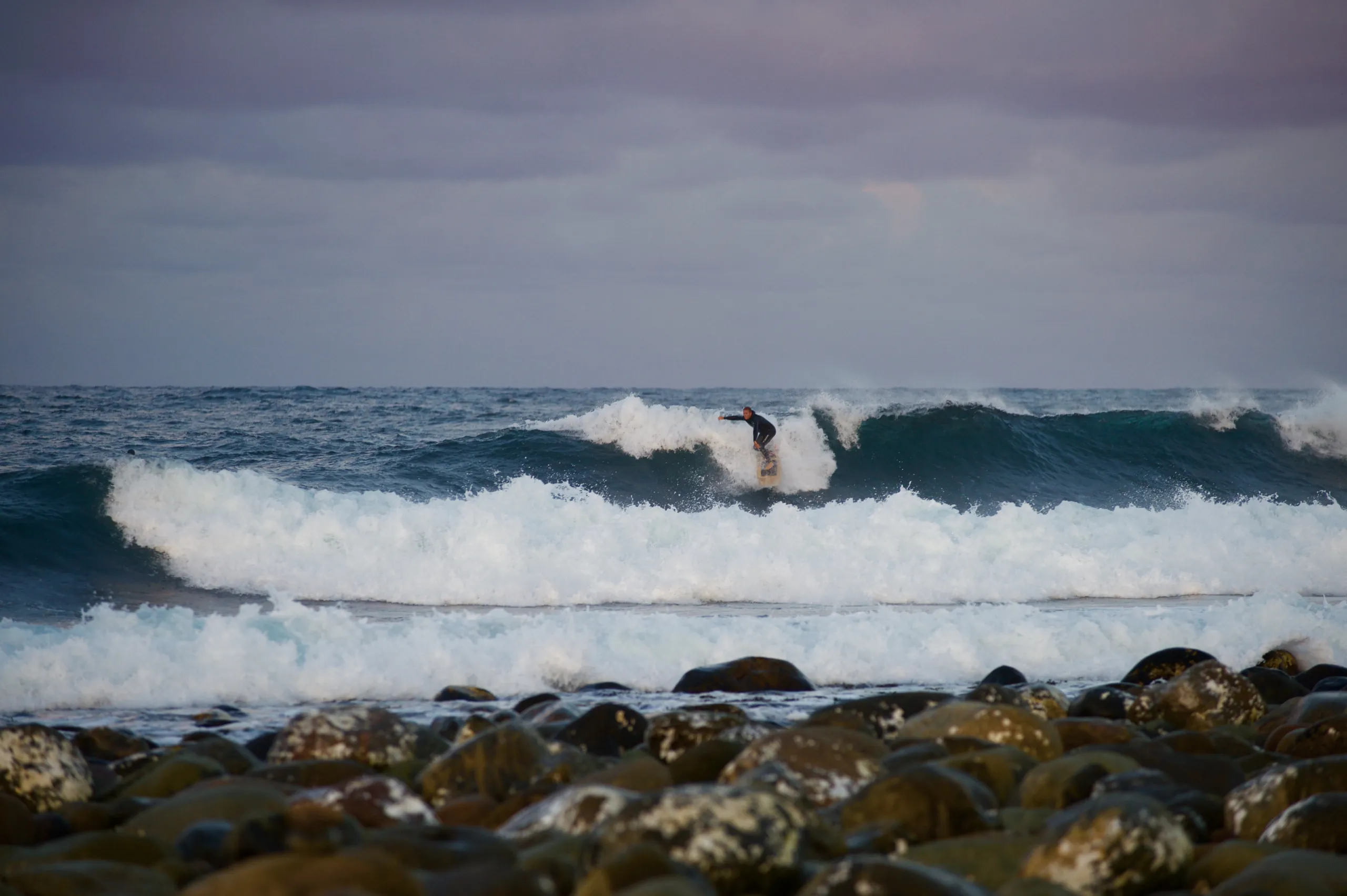 Surfer på en brytende bølge i skumringen, med rullesteiner i forgrunnen