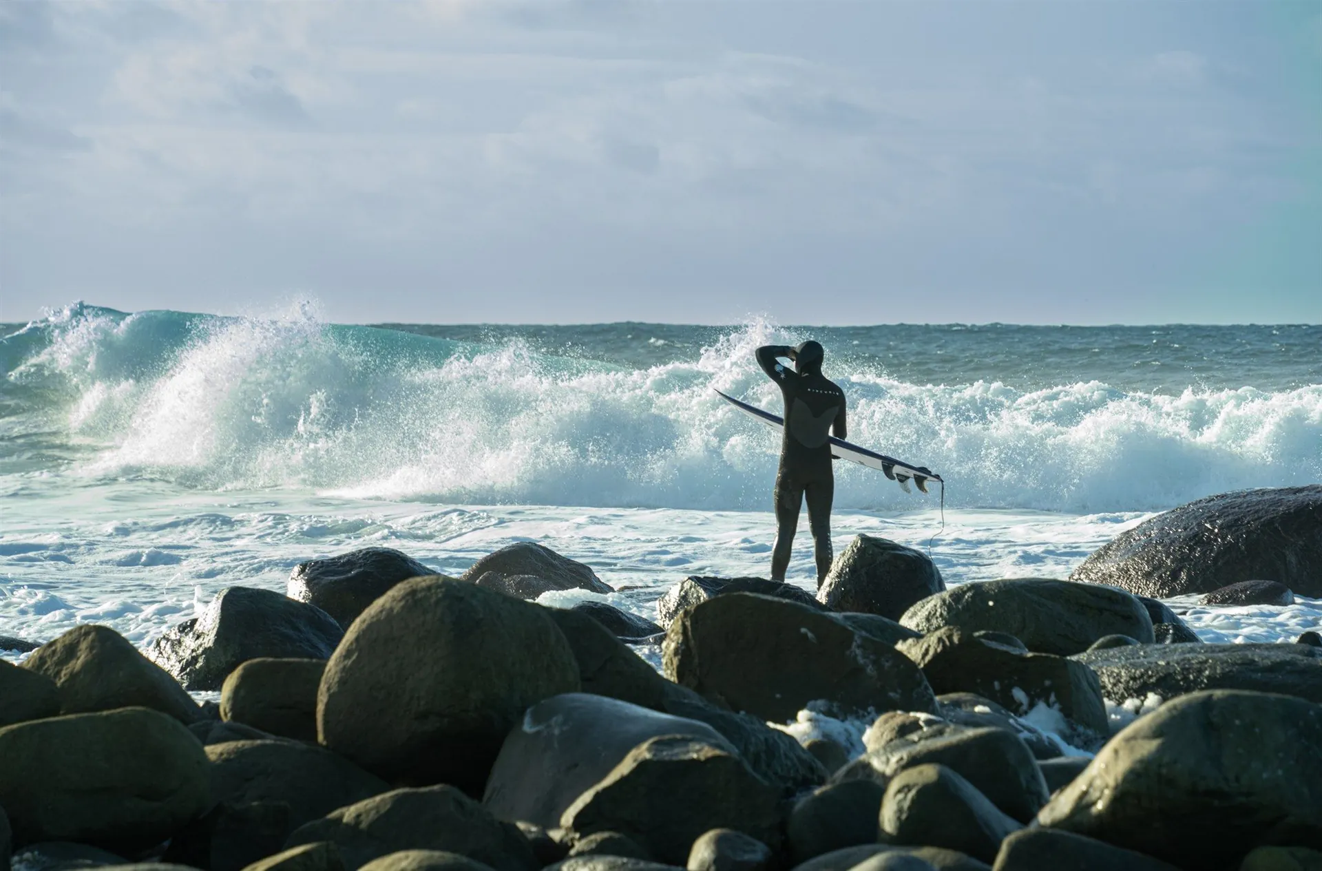 Surfer med brettet under armen står på rullesteinene ved Pisserenna og ser utover bølgene