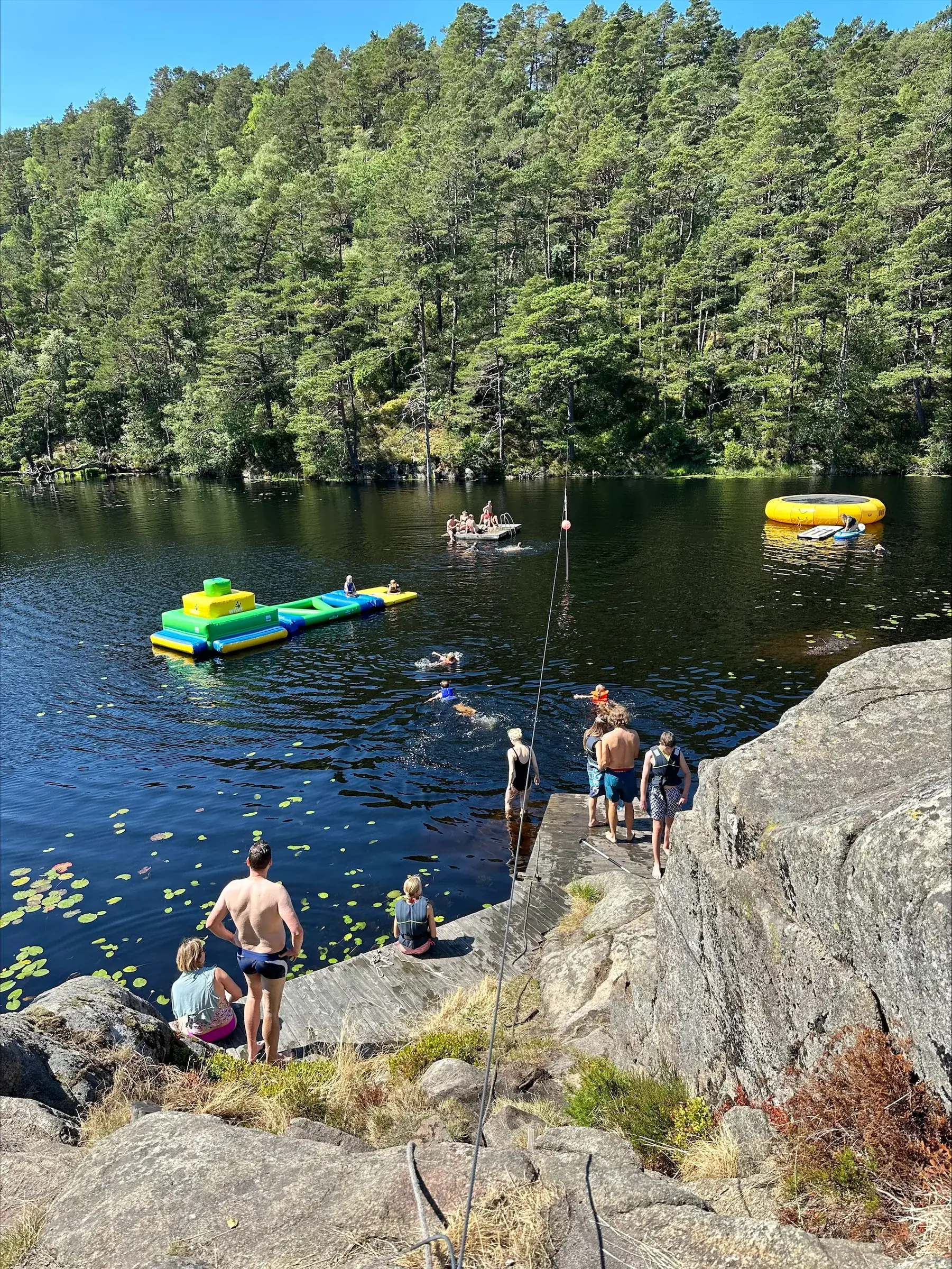 Badende ved svabergene i Engelsvannet, med flytebrygge og skog i bakgrunnen