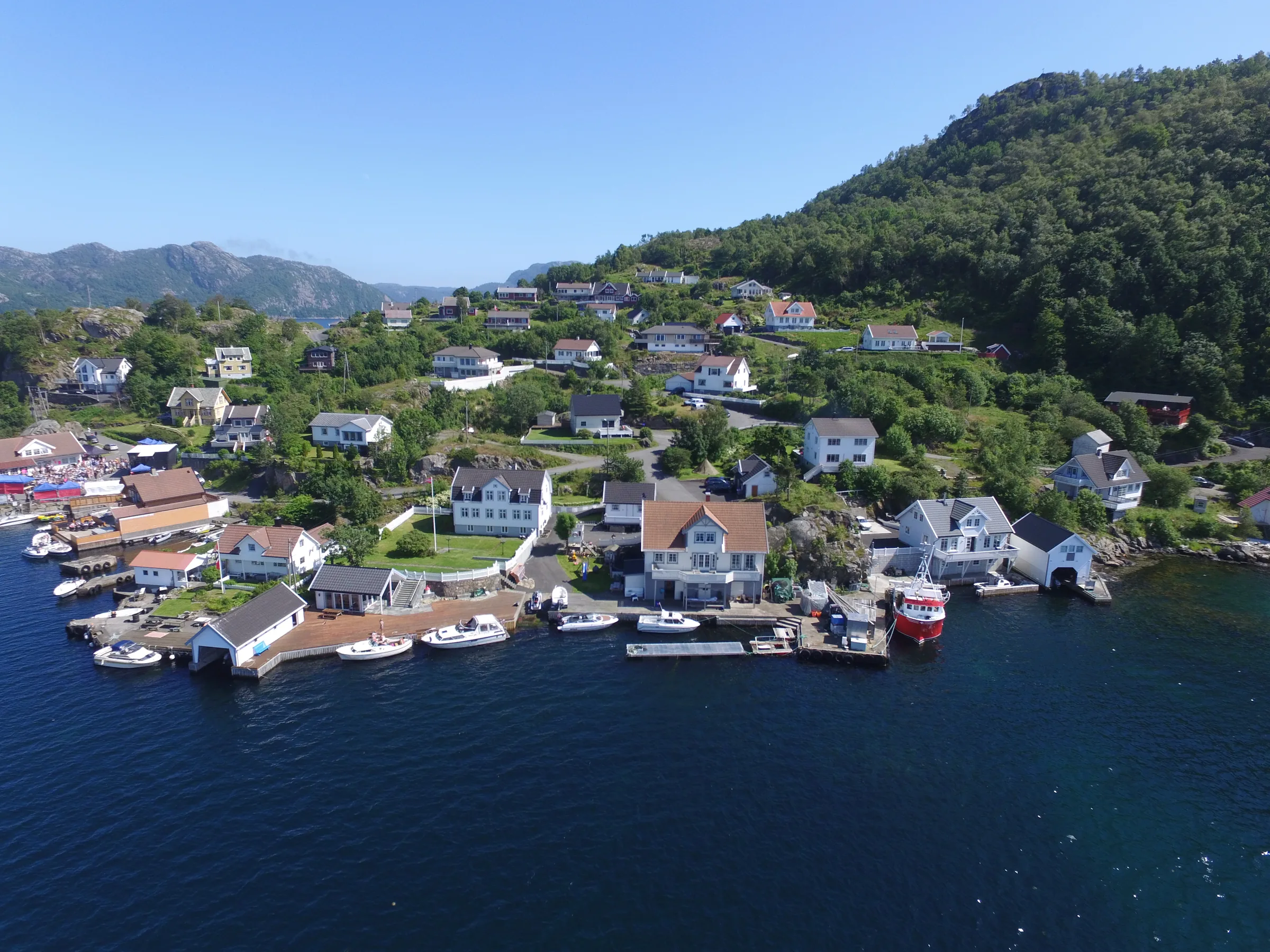 Flyfoto av bygda på Andabeløy: hvite hus og naust langs den lune fjorden, med skogkledde åser bak