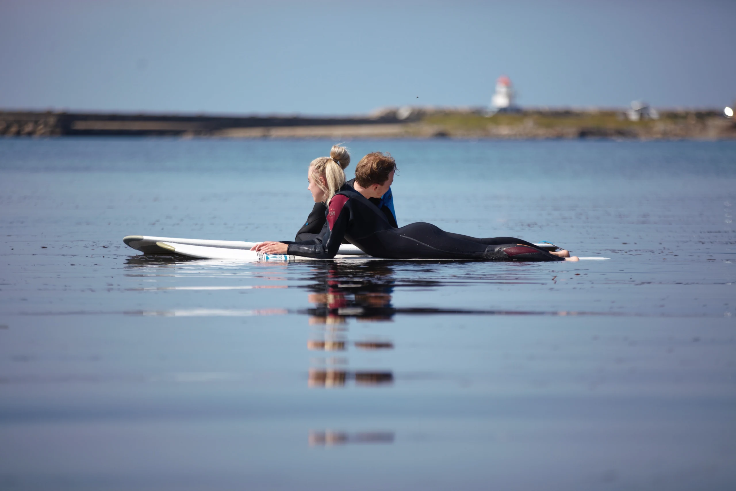 To personer i våtdrakt ligger på surfebrettene i blikkstille vann i solnedgang, med Brekneholmen fyrlykt i bakgrunnen
