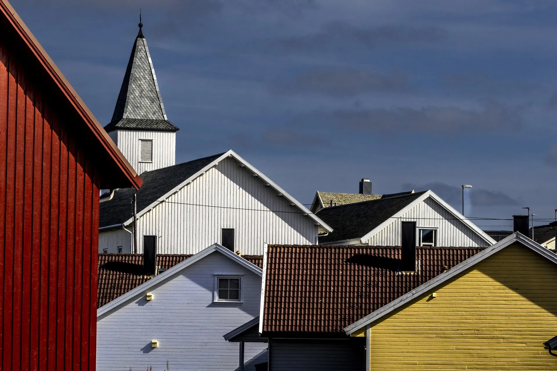 Kirketårn og hustak i hvitt, rødt og gult mot en mørk himmel på Borhaug