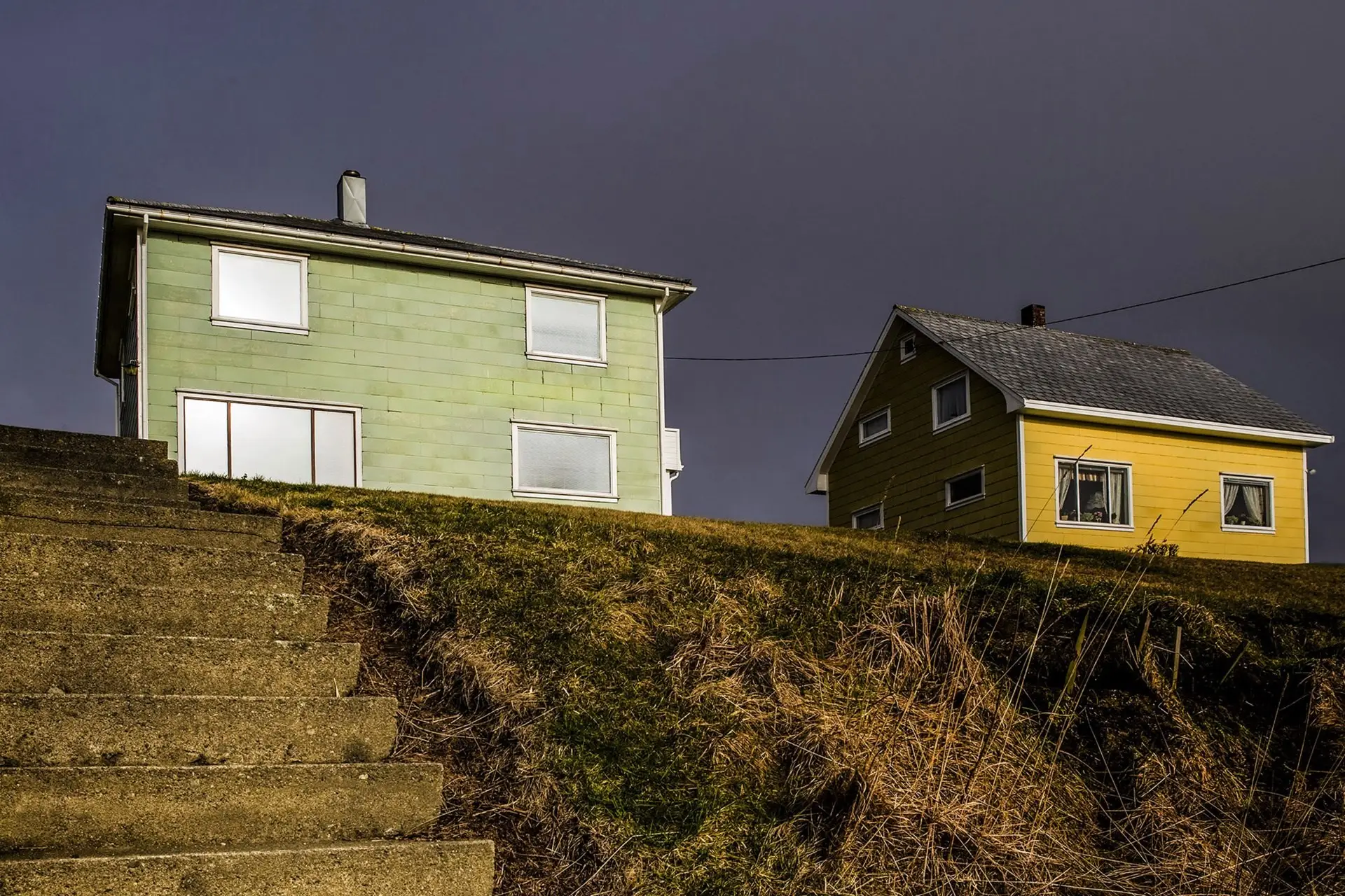 Et grønt og et gult trehus på en gresskledd kolle over en steintrapp, under mørk himmel