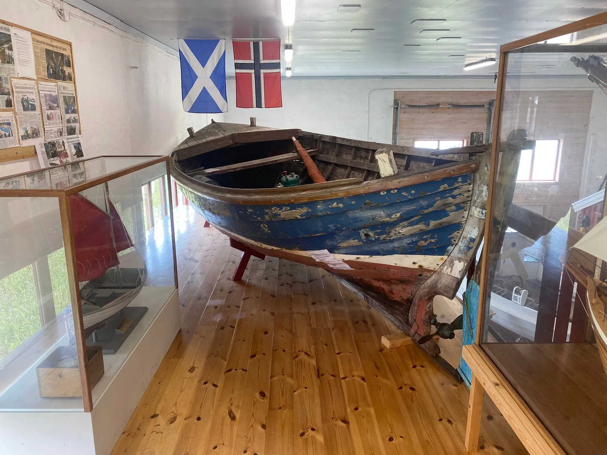 Åpen trebåt med blå skrog utstilt innendørs i kystkultursenteret, med flagg og fotografier på veggen bak