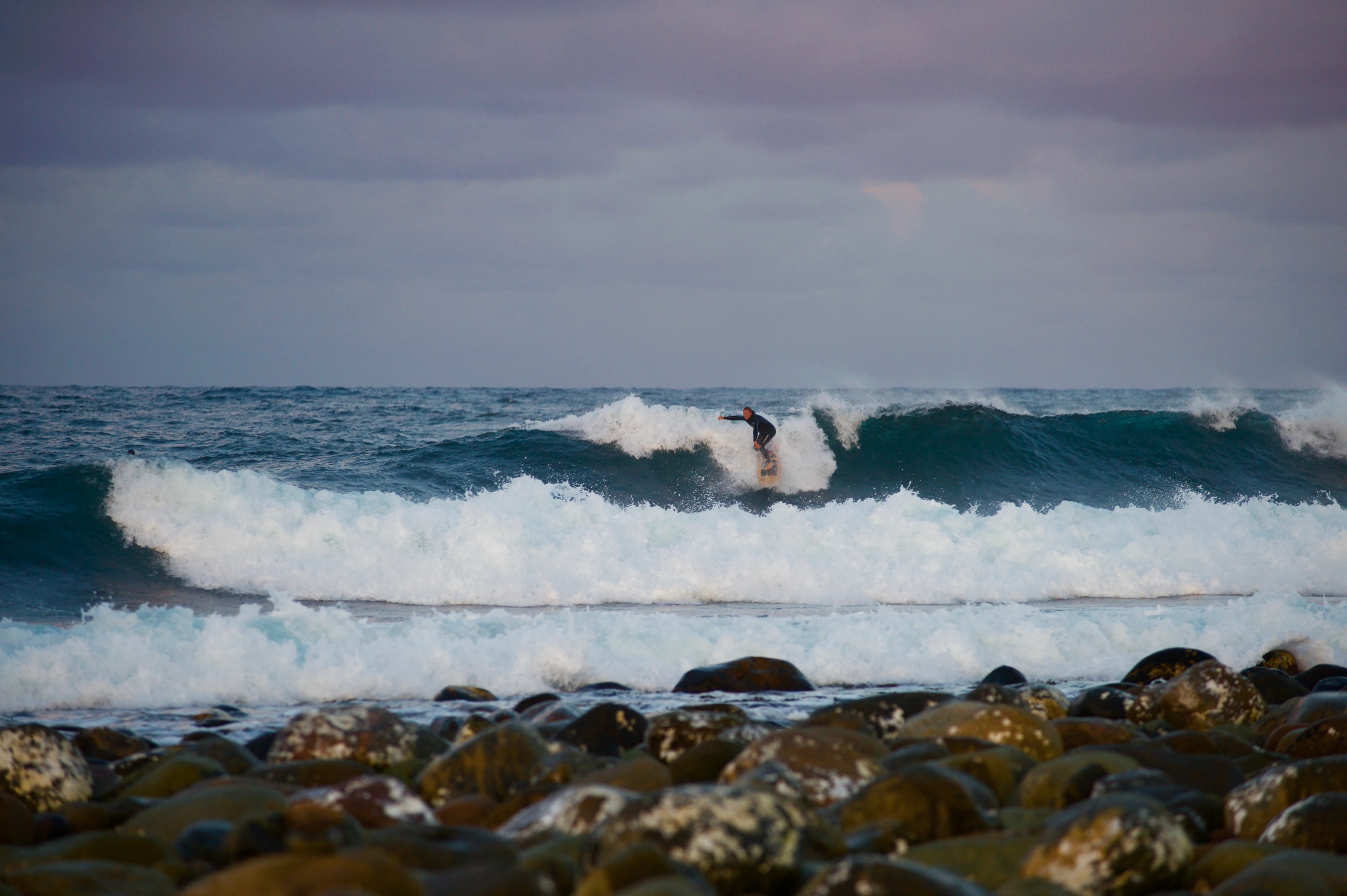 Surfer rir en bølge inn mot rullesteinene på Lista, med mørk himmel over havet