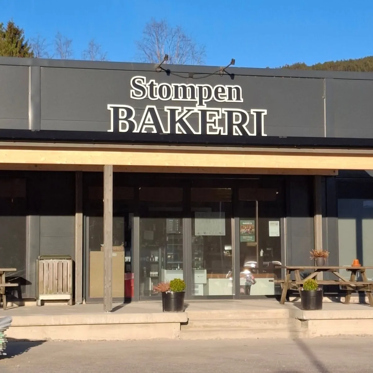 Fasaden til Stompen bakeri med skiltet over inngangen, benker og blomsterkrukker foran, under blå himmel