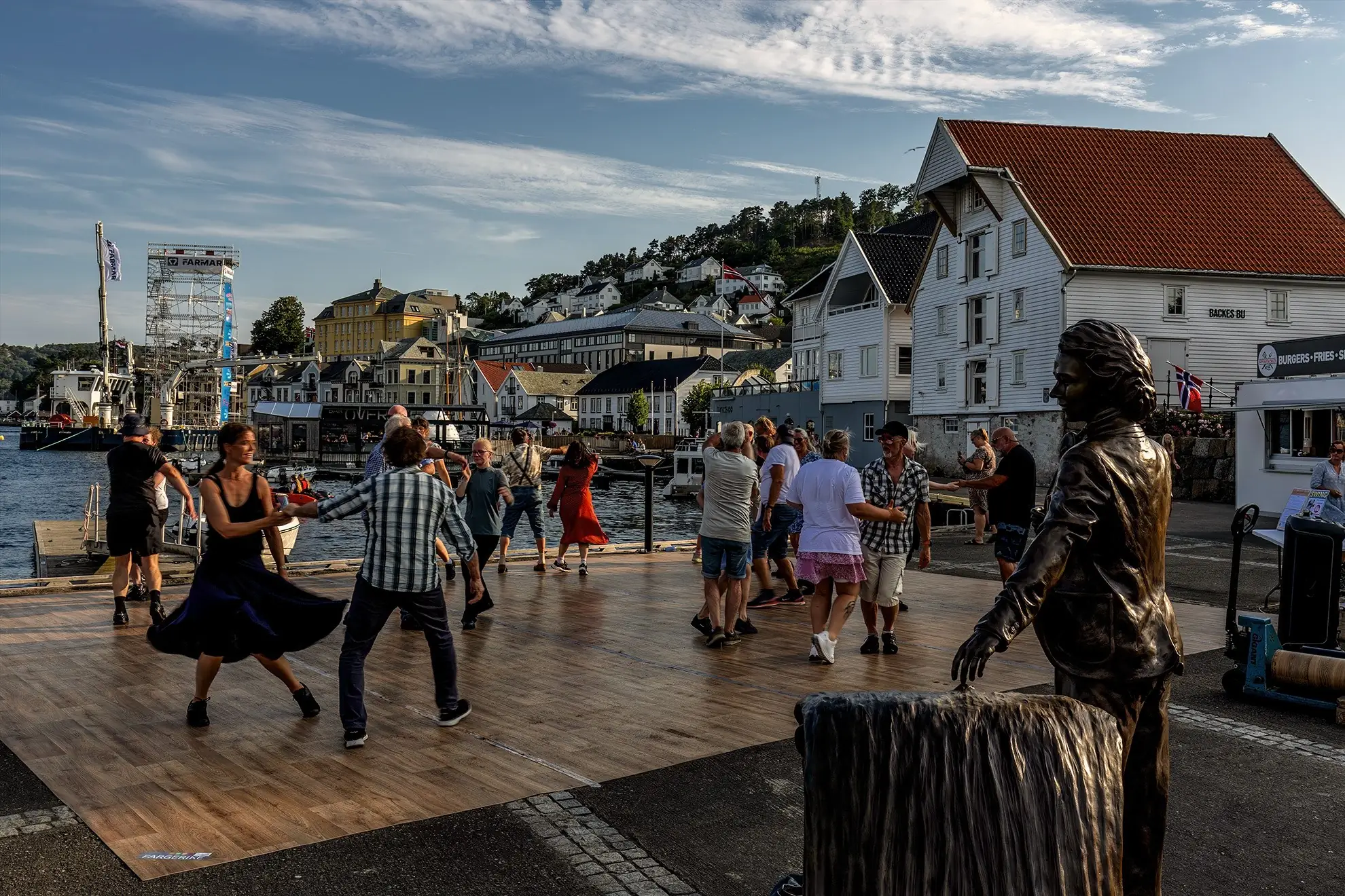 Folk som danser på en dansebane ved havna på Fern Sundes plass, med en bronsestatue i forgrunnen og hvite hus bak