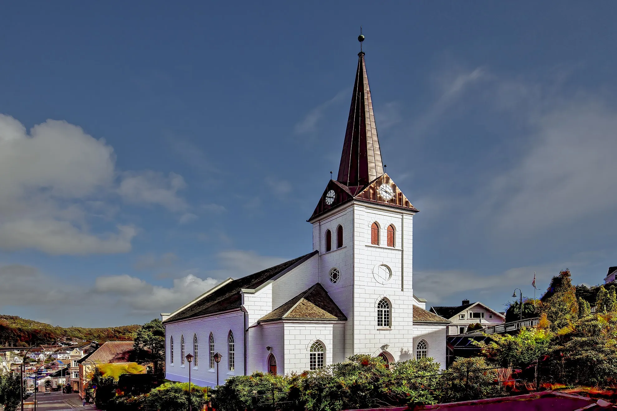 Frelserens kirke i Farsund, hvit med mørkt spir, en solrik dag med blå himmel og hustak nedenfor