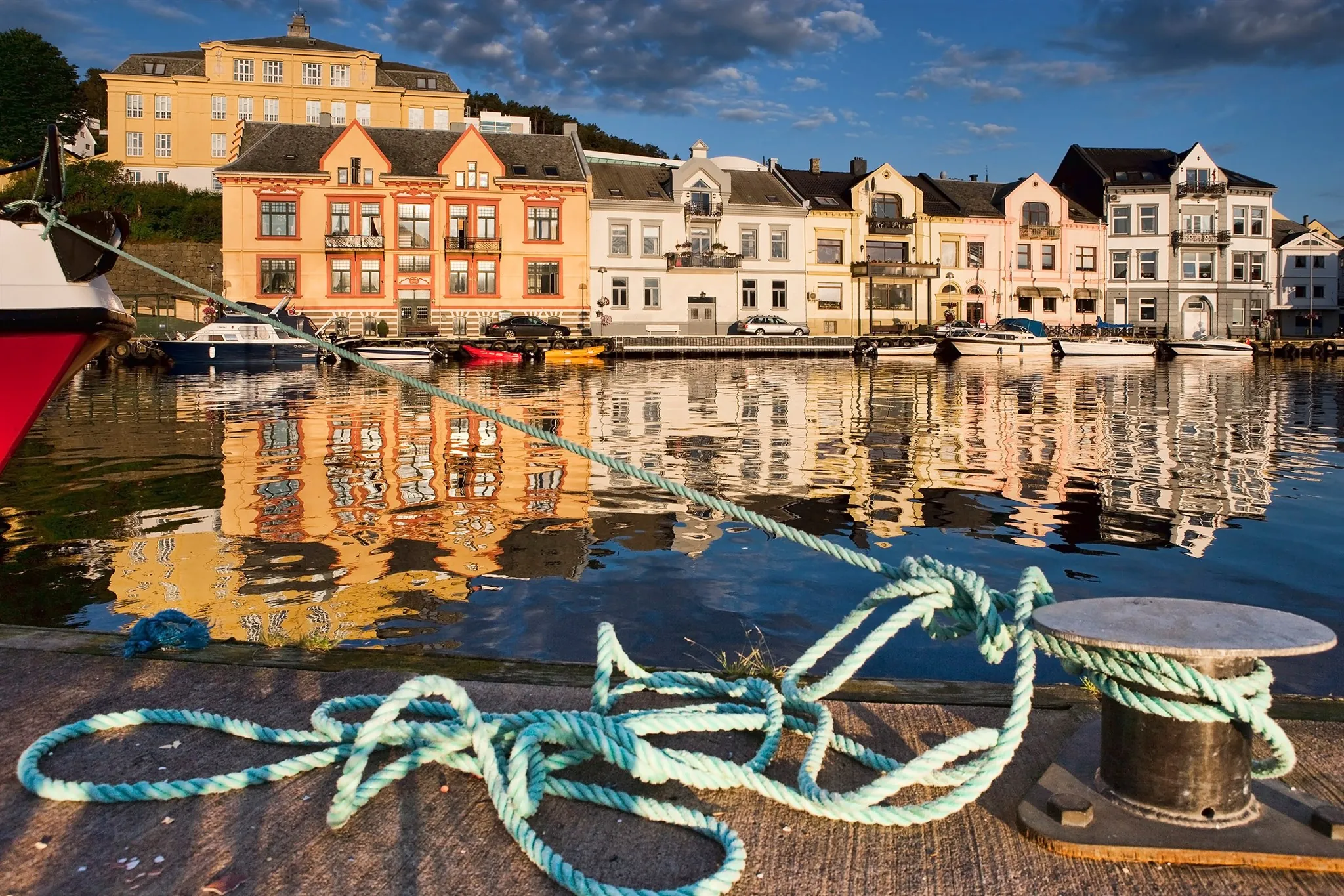 Fargerike bygårder langs havna i Farsund i kveldssol, speilet i vannet, med et tau rundt en pullert på kaia i forgrunnen