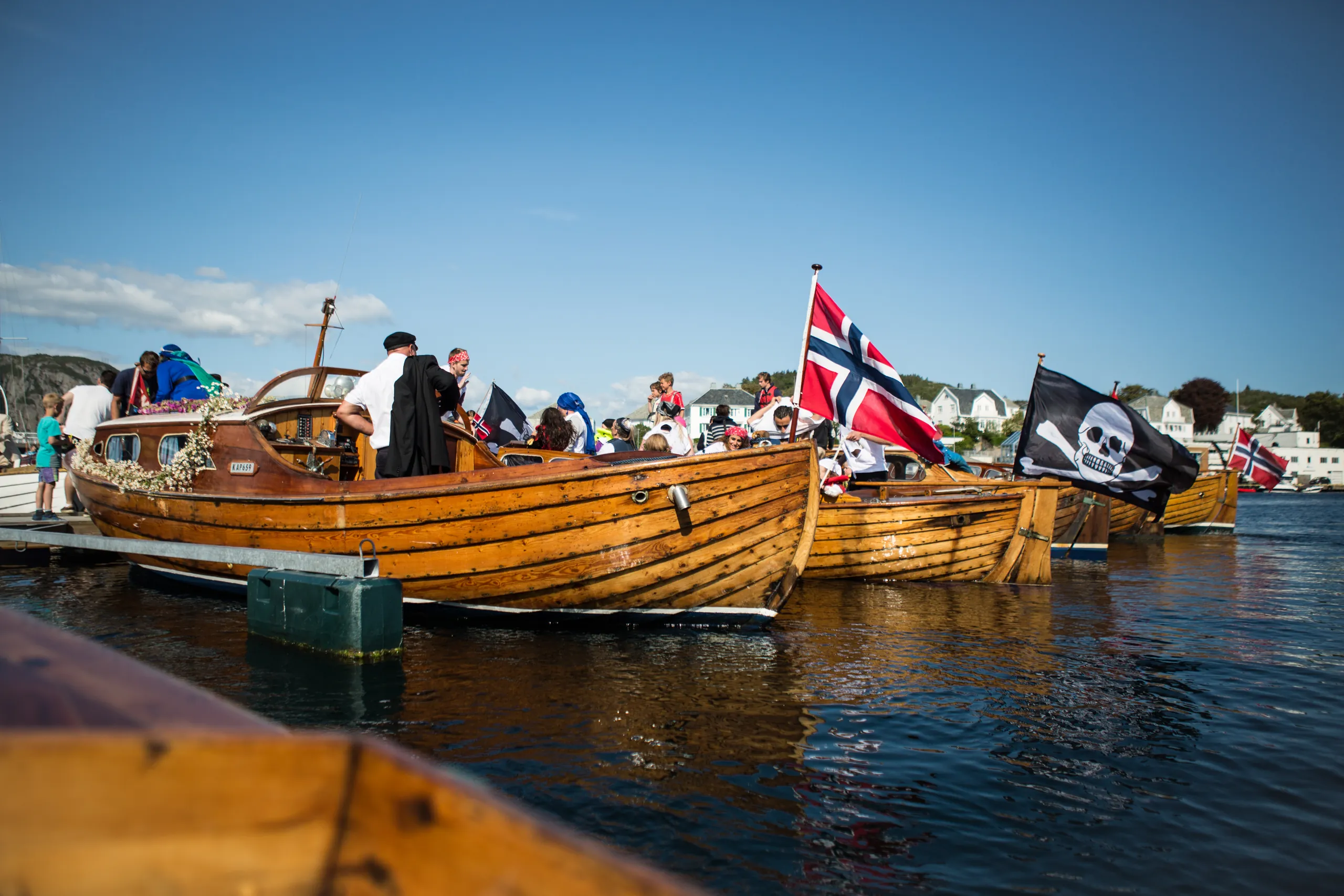 Trebåter pyntet med norske flagg og sjørøverflagg ligger side om side i havna under Kaperdagene, med folk i kostymer om bord