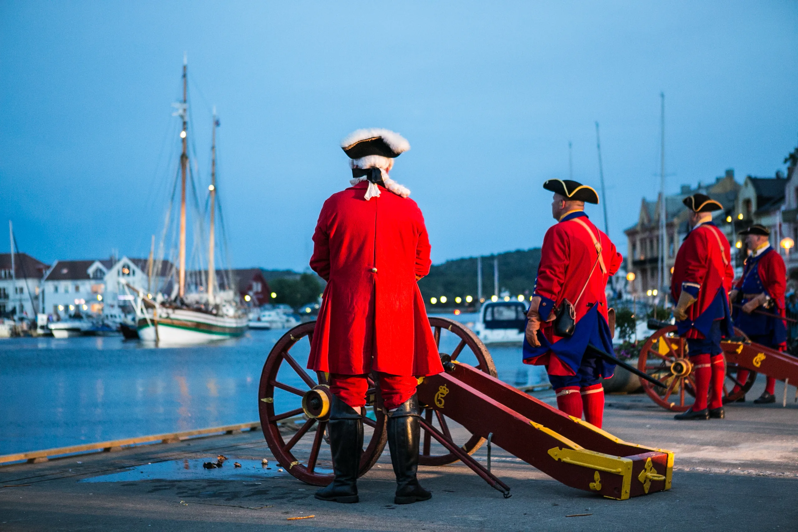 Menn i røde kaperuniformer og trekantede hatter ved en kanon på kaia i Farsund i blåtimen, seilskuter i havna bak