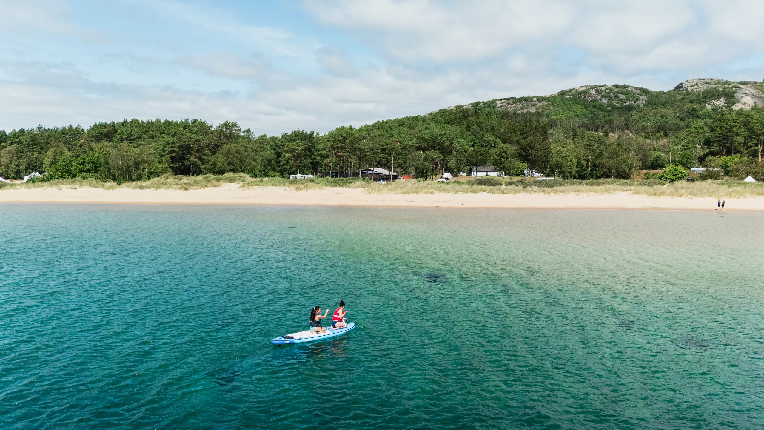 Lomsesanden fra lufta: hvit strand, turkist langgrunt vann og en padlebrett-padler med barn, campingplassen i skogkanten