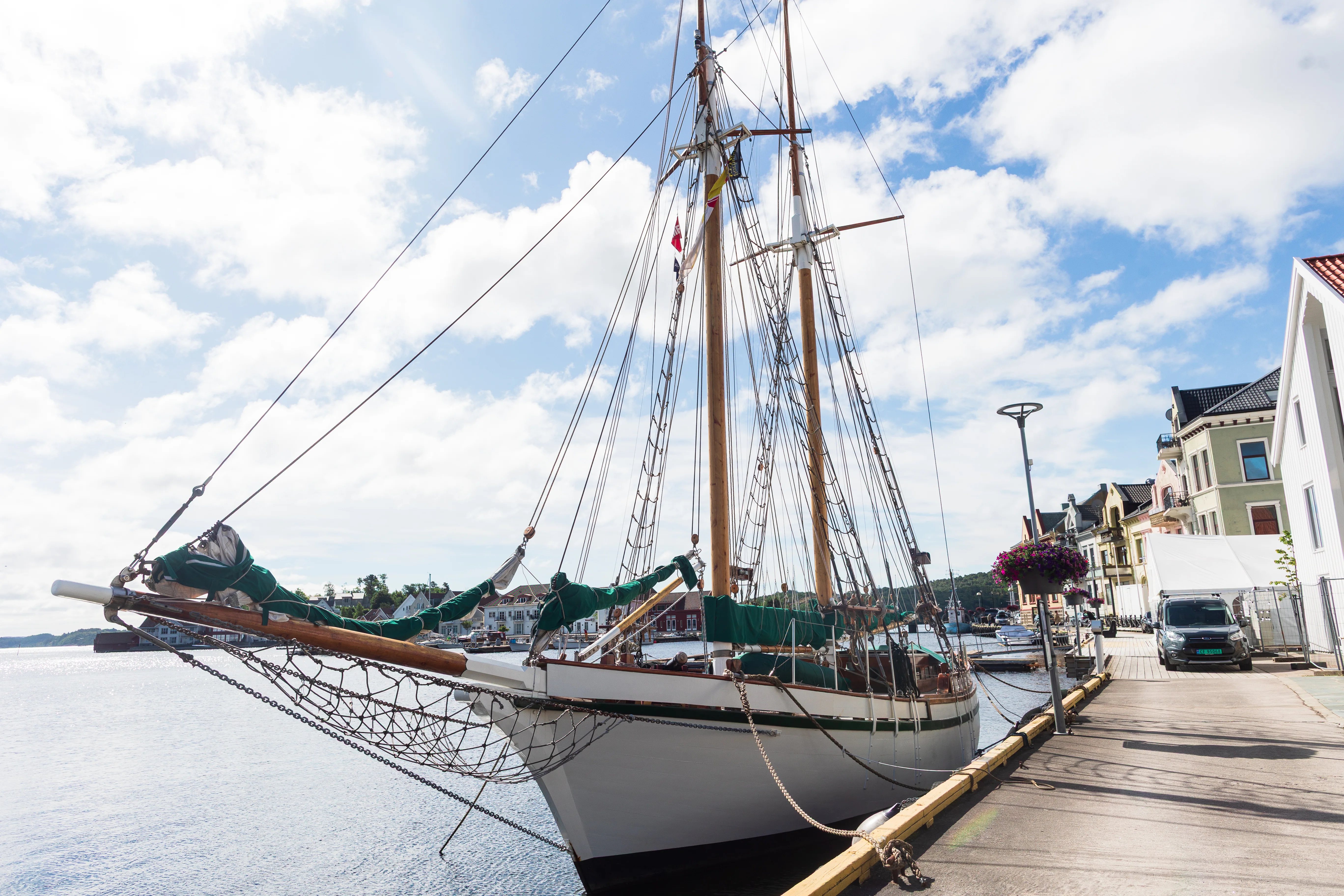 Tomastet seilskute fortøyd ved kaia i Farsund sentrum en sommerdag, med bygårdene langs havna til høyre