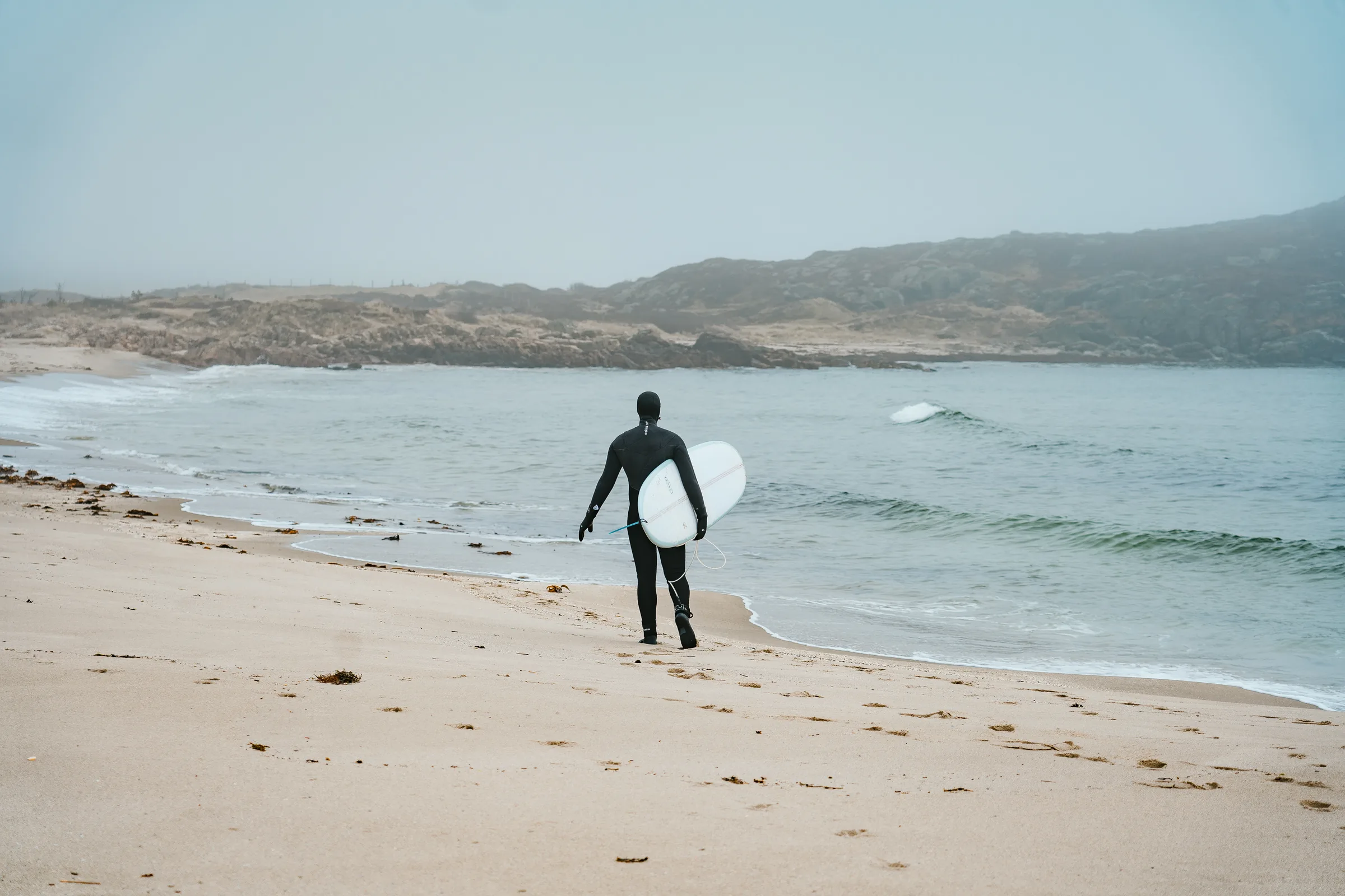Surfer i våtdrakt går med brettet under armen langs vannkanten på Haviksanden, med bølgene og klippene bak