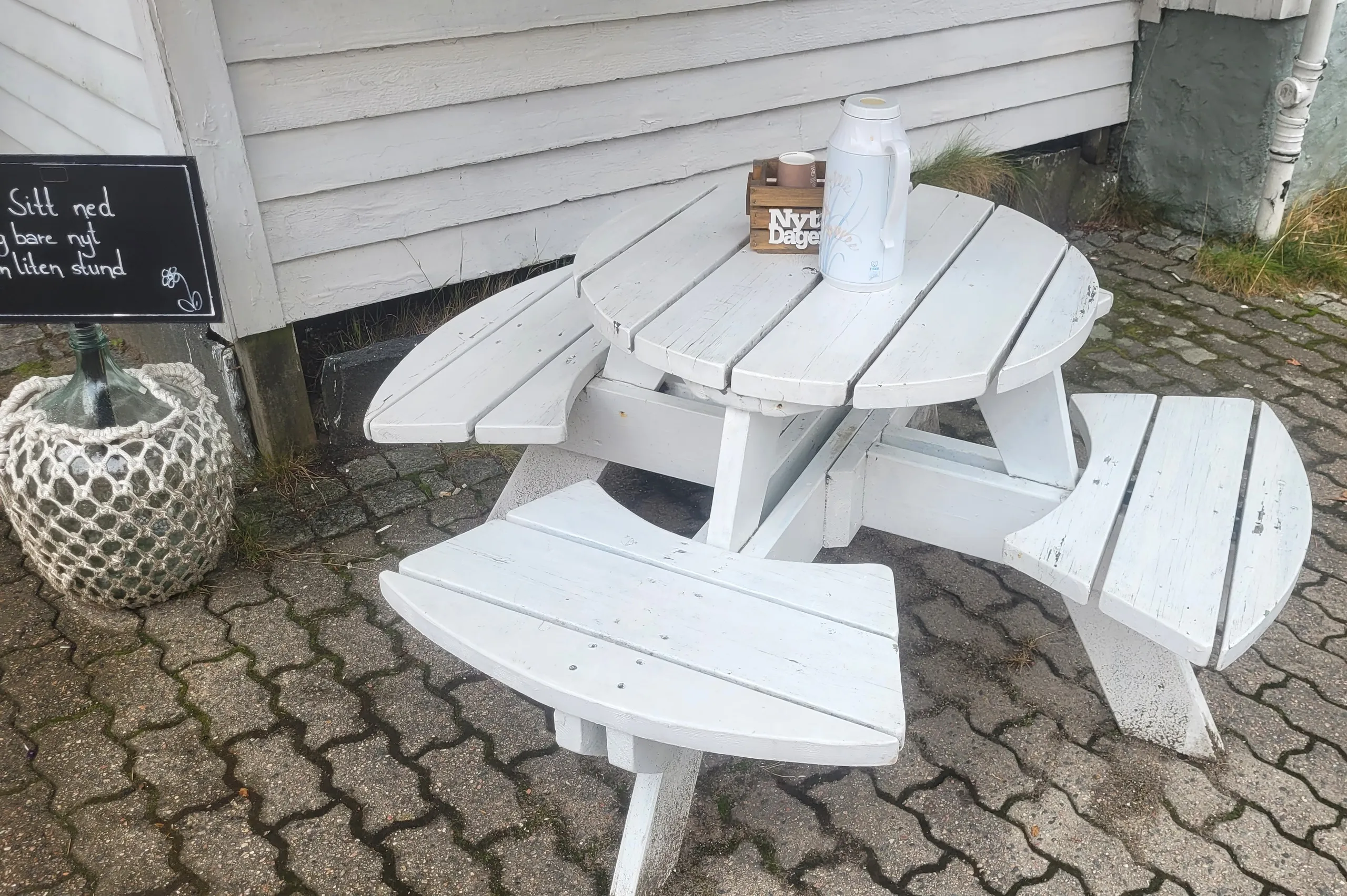 Et lite hvitmalt bord med termos og kopper på brosteinen, og en tavle med invitasjon til gratis kaffe