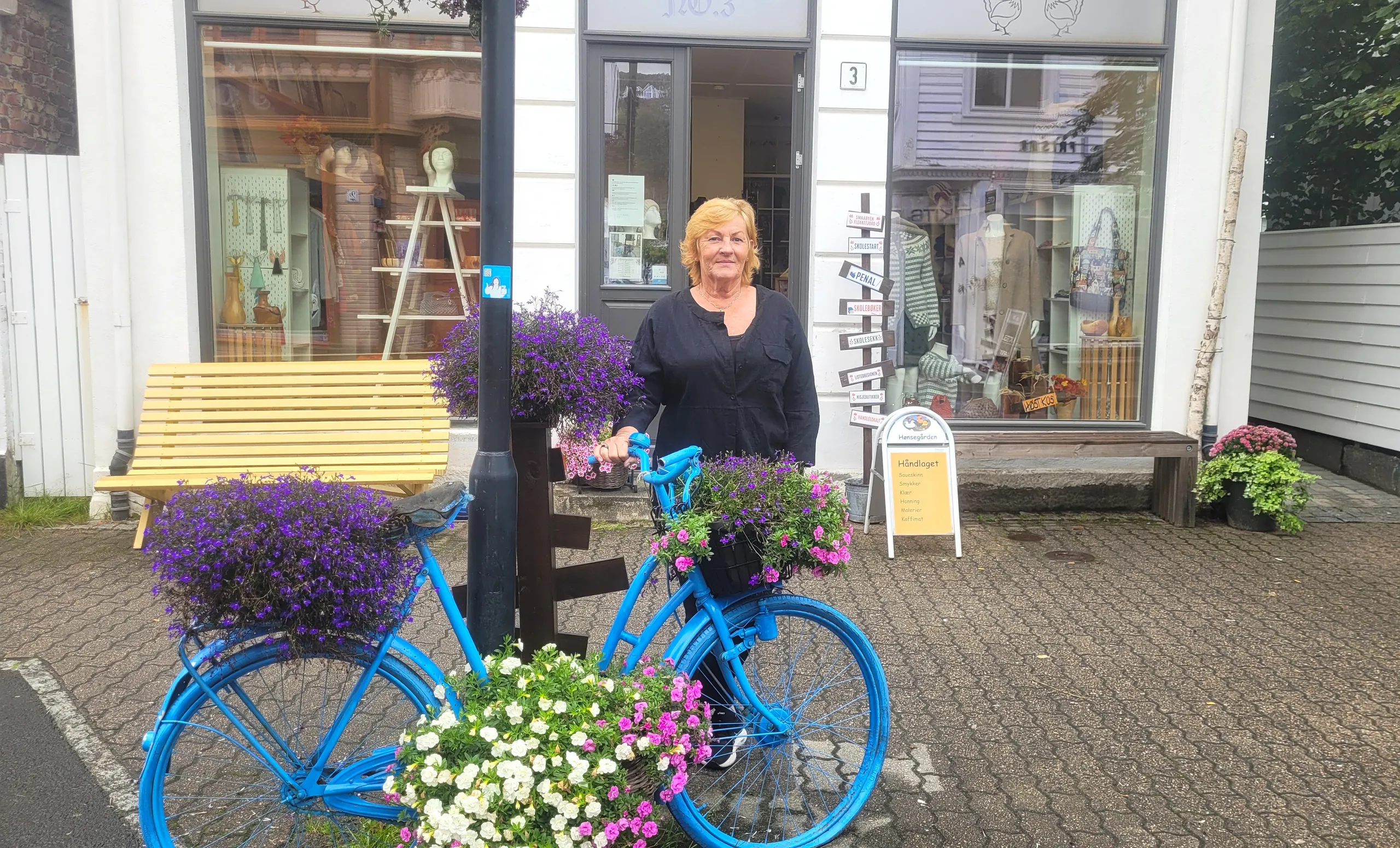 Mette Syvertsen står med en blå sykkel full av lilla blomster utenfor butikken Hønsegården i Flekkefjord