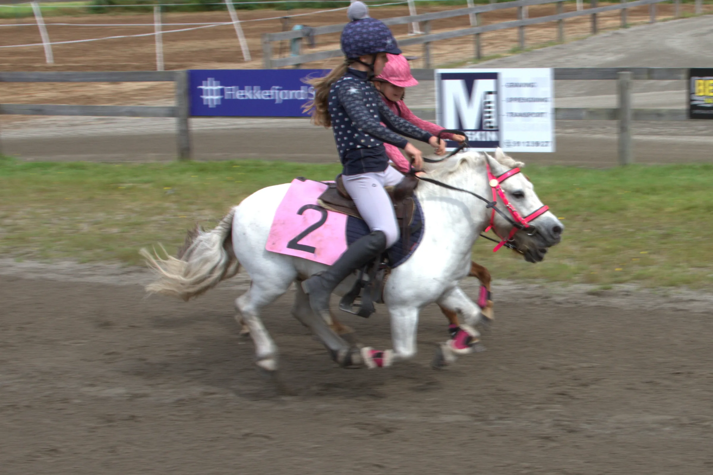 To jenter i hjelm rir side om side i galopp på grusbanen, den nærmeste på en hvit ponni med startnummer 2 og den andre på en brun ponni bak