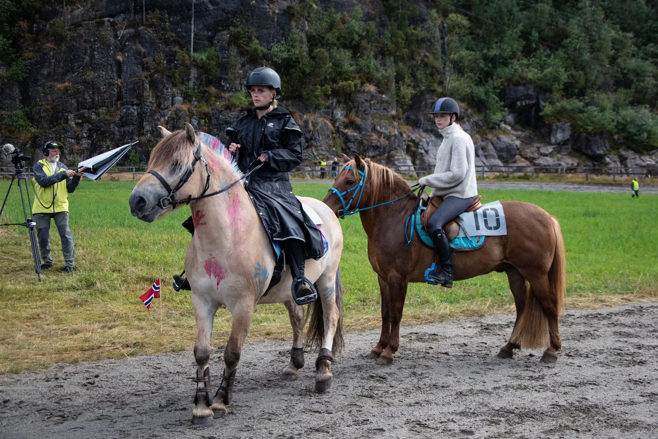 To unge ryttere i hjelm på hver sin hest på banen, den lyse hesten med rosa og blå malte flekker og farget man, mens en mann filmer fra kanten foran fjellveggen