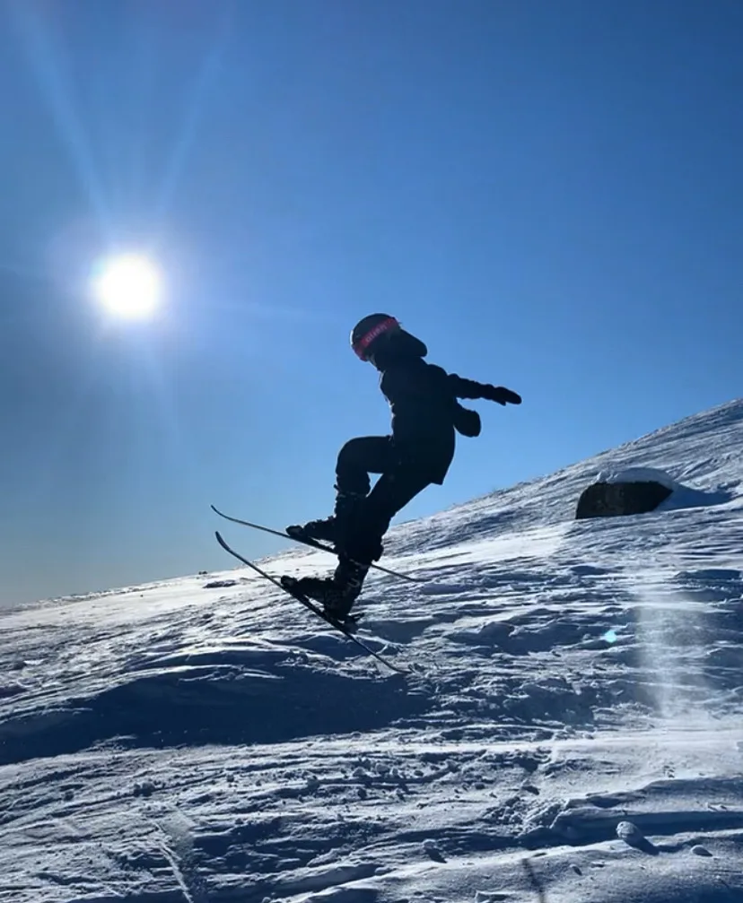 En snowboardkjører i silhuett tar et hopp i snøen, med sola høyt på en dypblå vinterhimmel