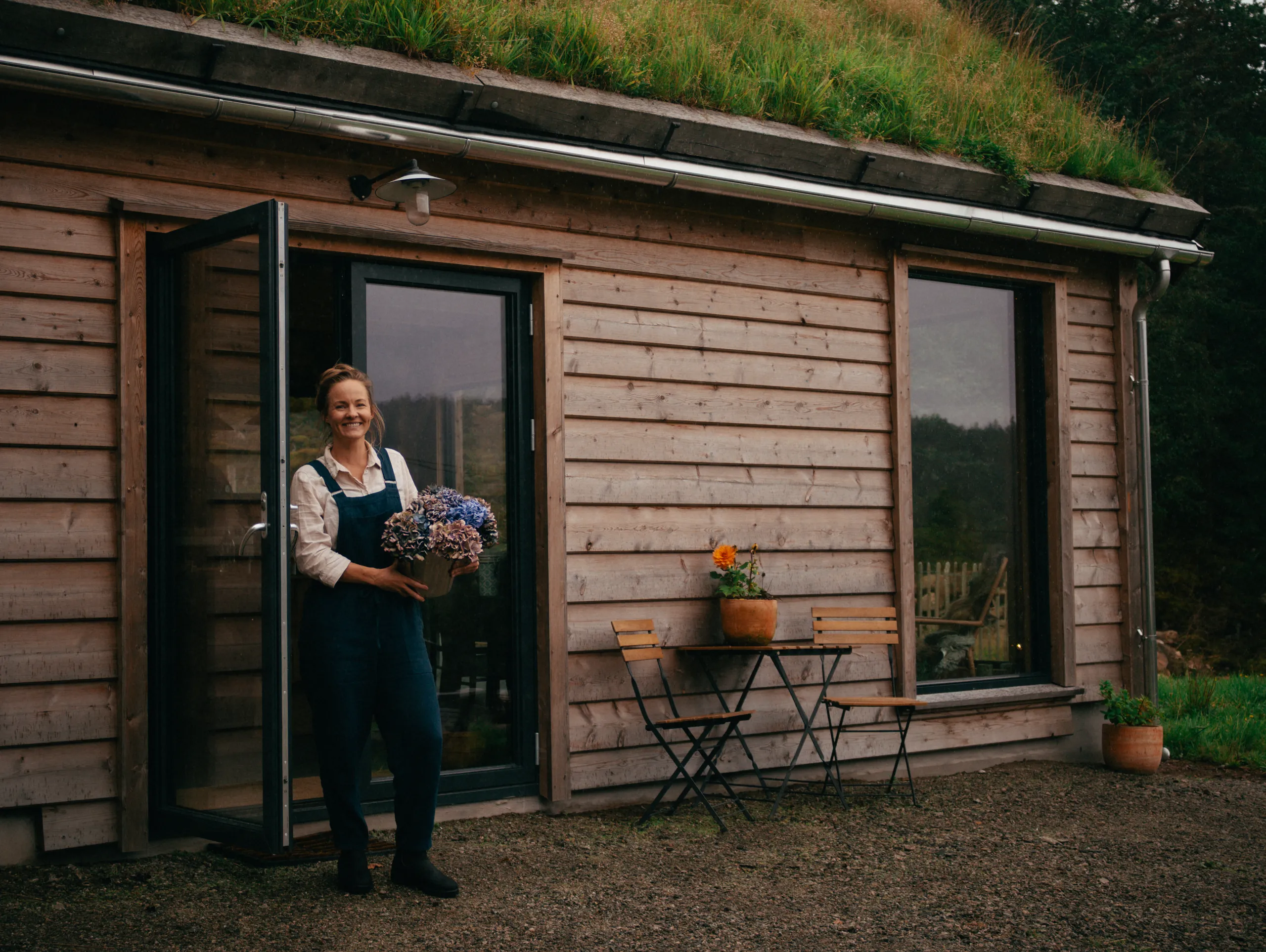 Nina står i døra til et grindbygg i tre med torvtak, med en bukett hortensia i armene og et lite kafébord ved siden av