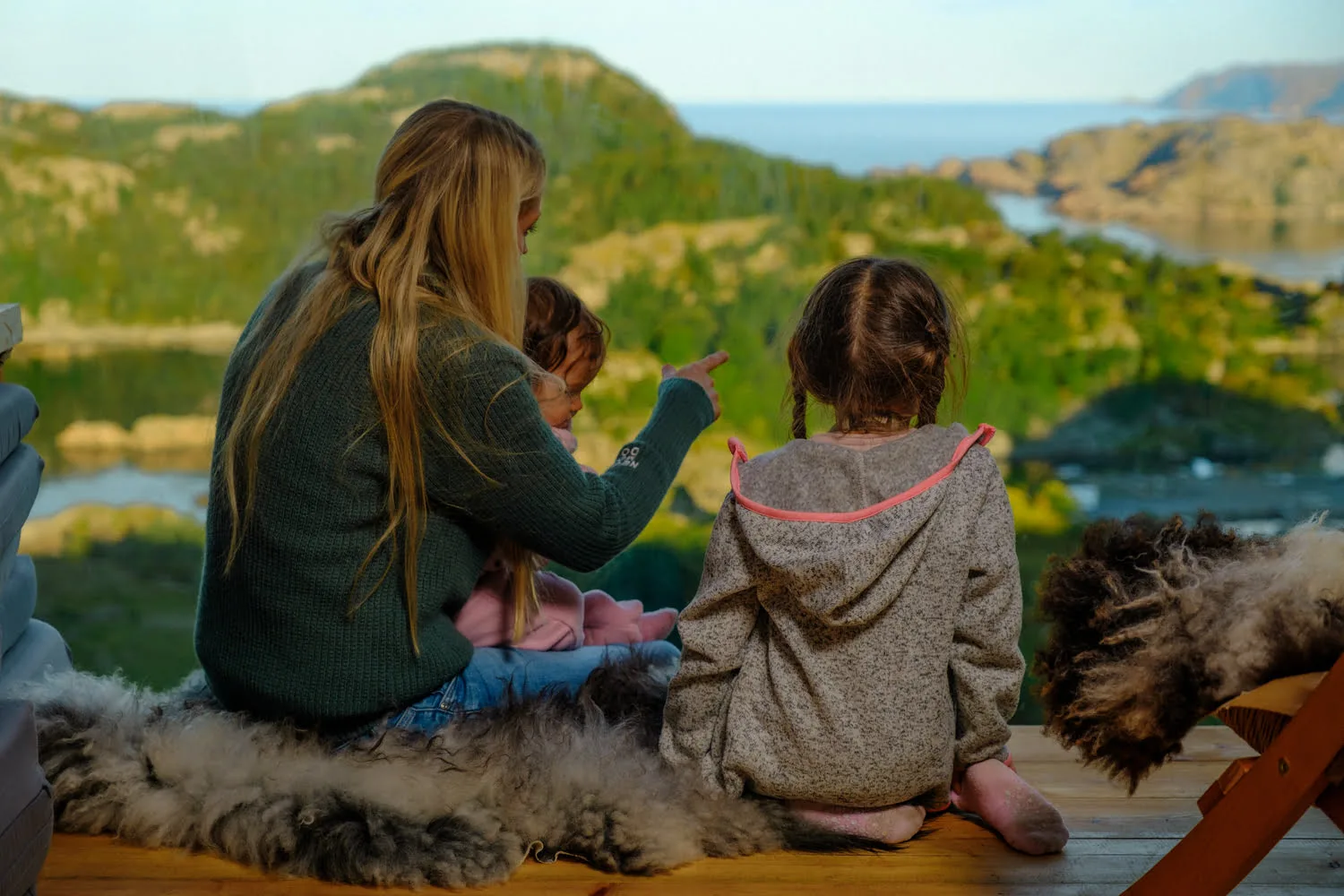 En mor og to barn sitter på saueskinn på en fjelltopp og ser ut over havet ved Hidra