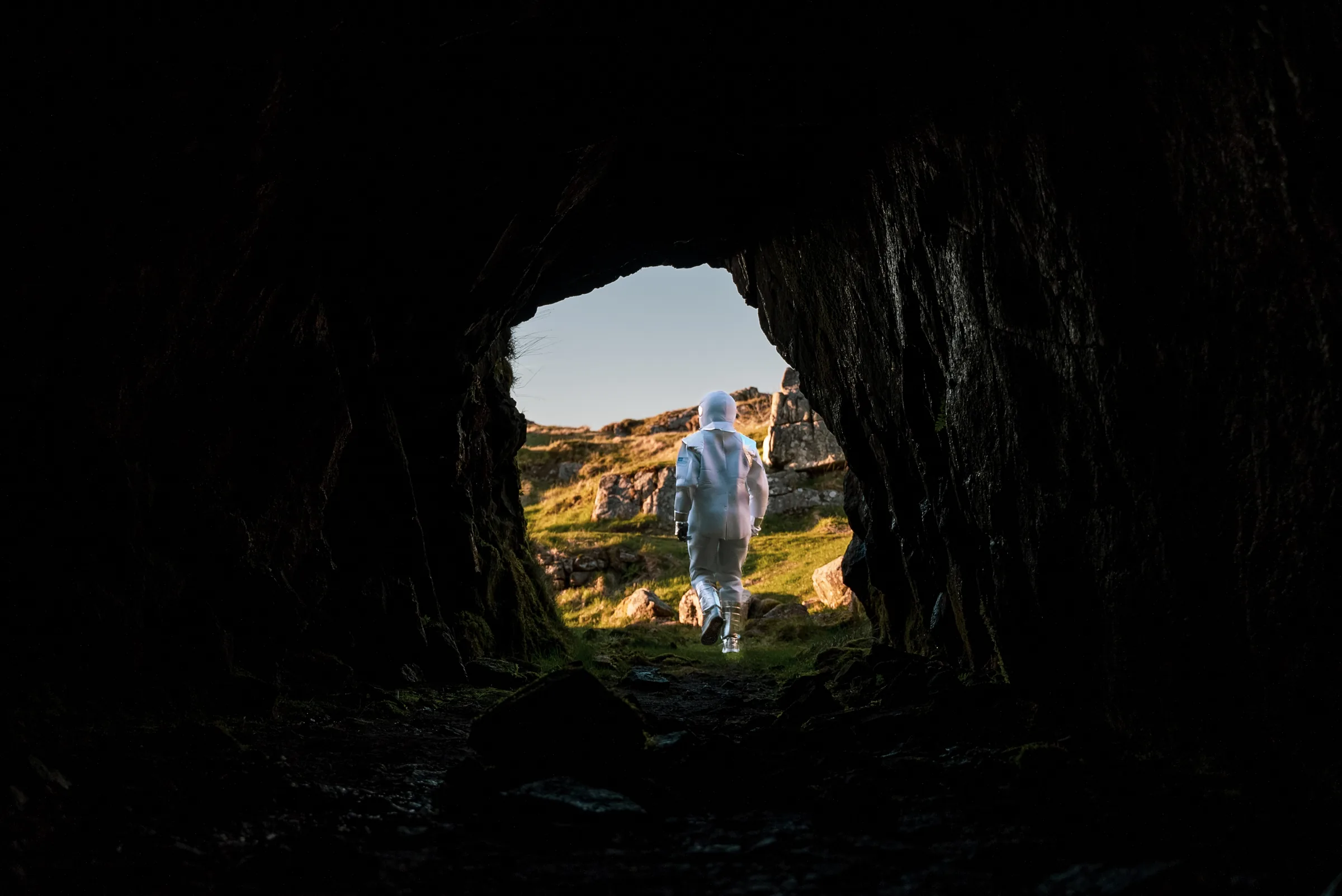 En person i astronautdrakt sett gjennom åpningen i en mørk hule, med lyst fjellandskap utenfor