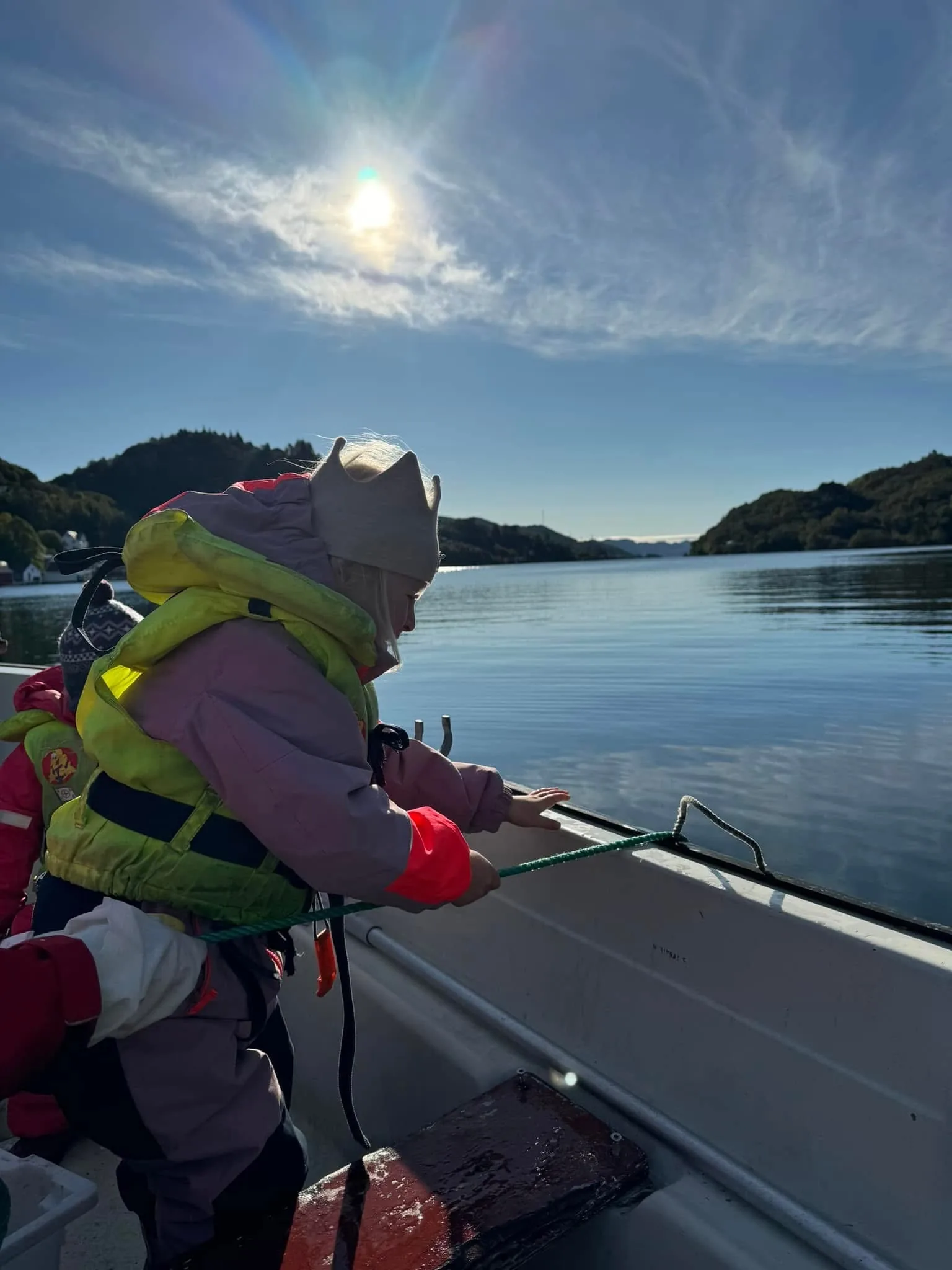Barnehagebarn i redningsvest i baugen på en småbåt, med lav sol over fjorden