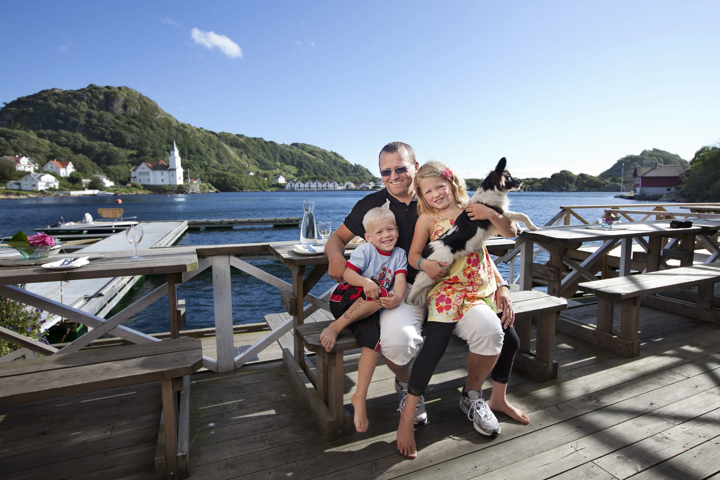 En familie med hund på uteserveringen til Isbua ved sjøkanten i Kirkehamn, i sol