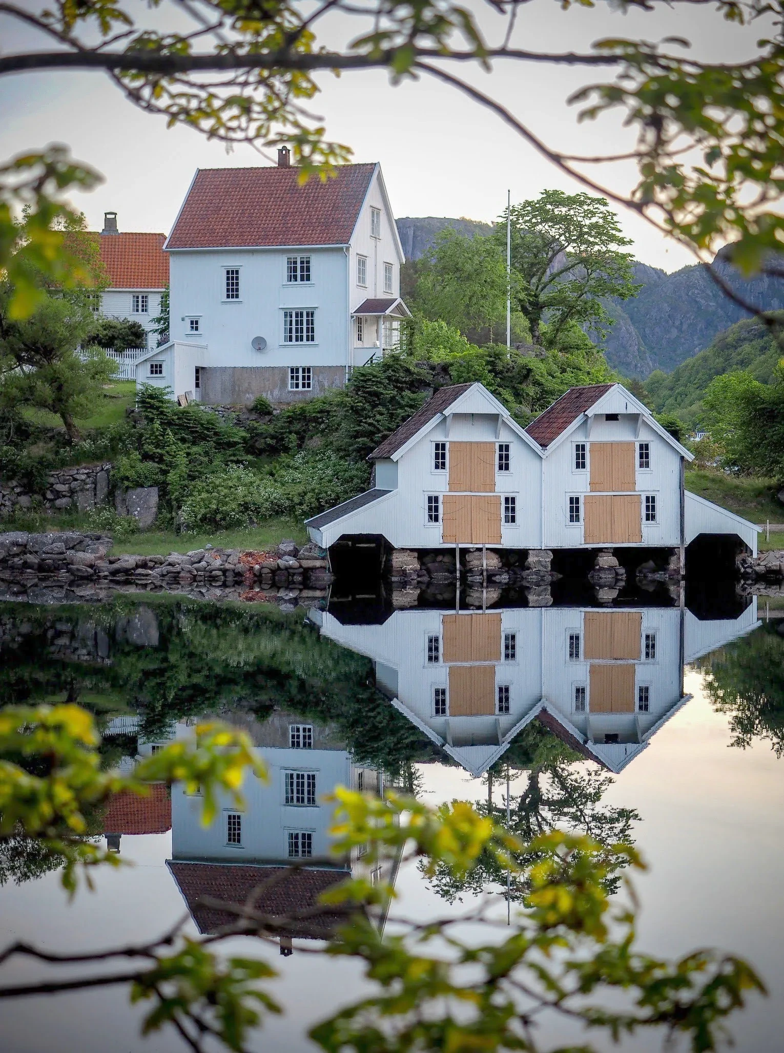 Hvitt trehus og to naust som speiler seg i blikkstille vann på Hidra